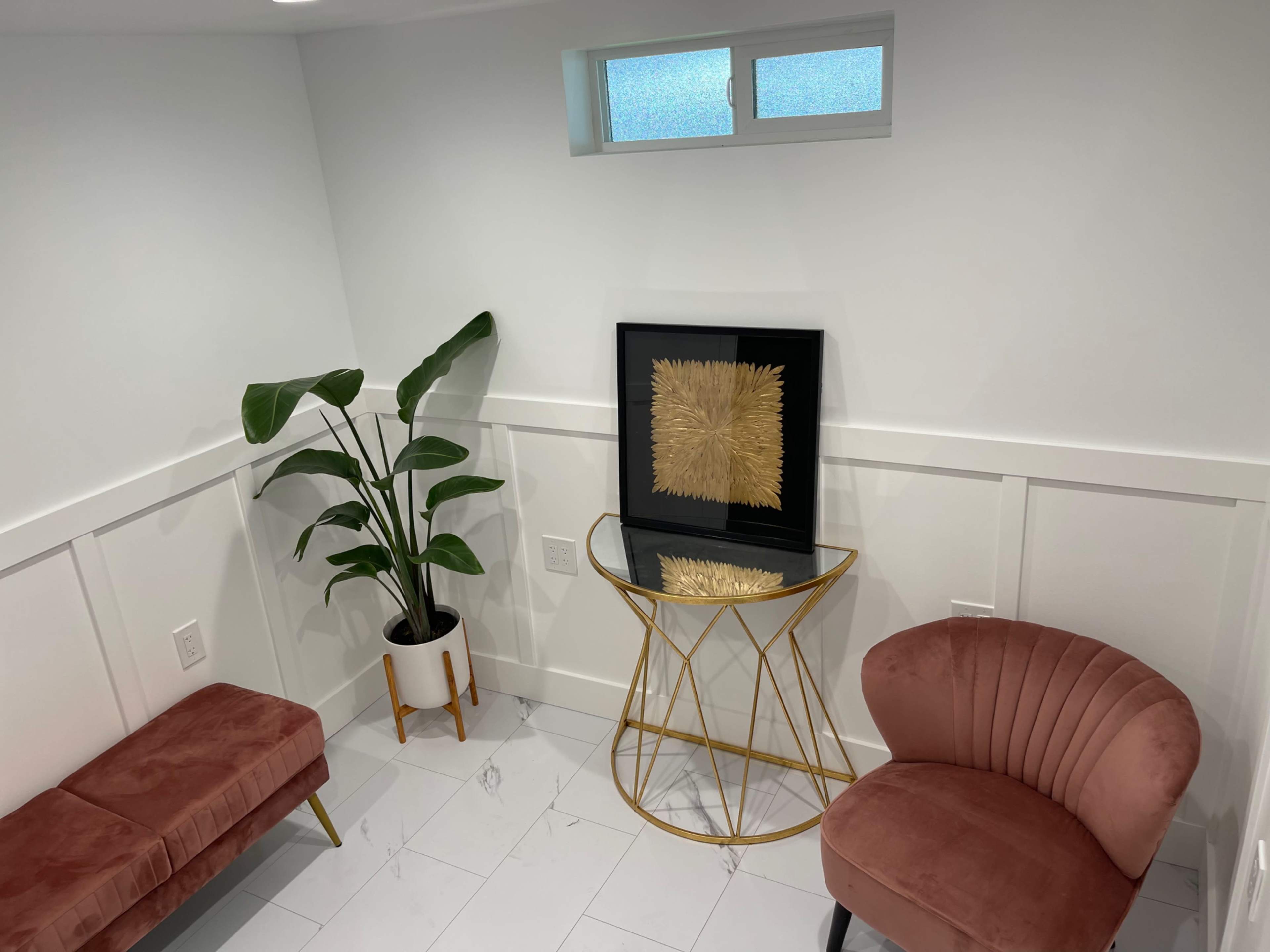 A small, well-lit room with a plant in a pot, a framed artwork on a gold side table, a pink upholstered chair, and a pink bench.