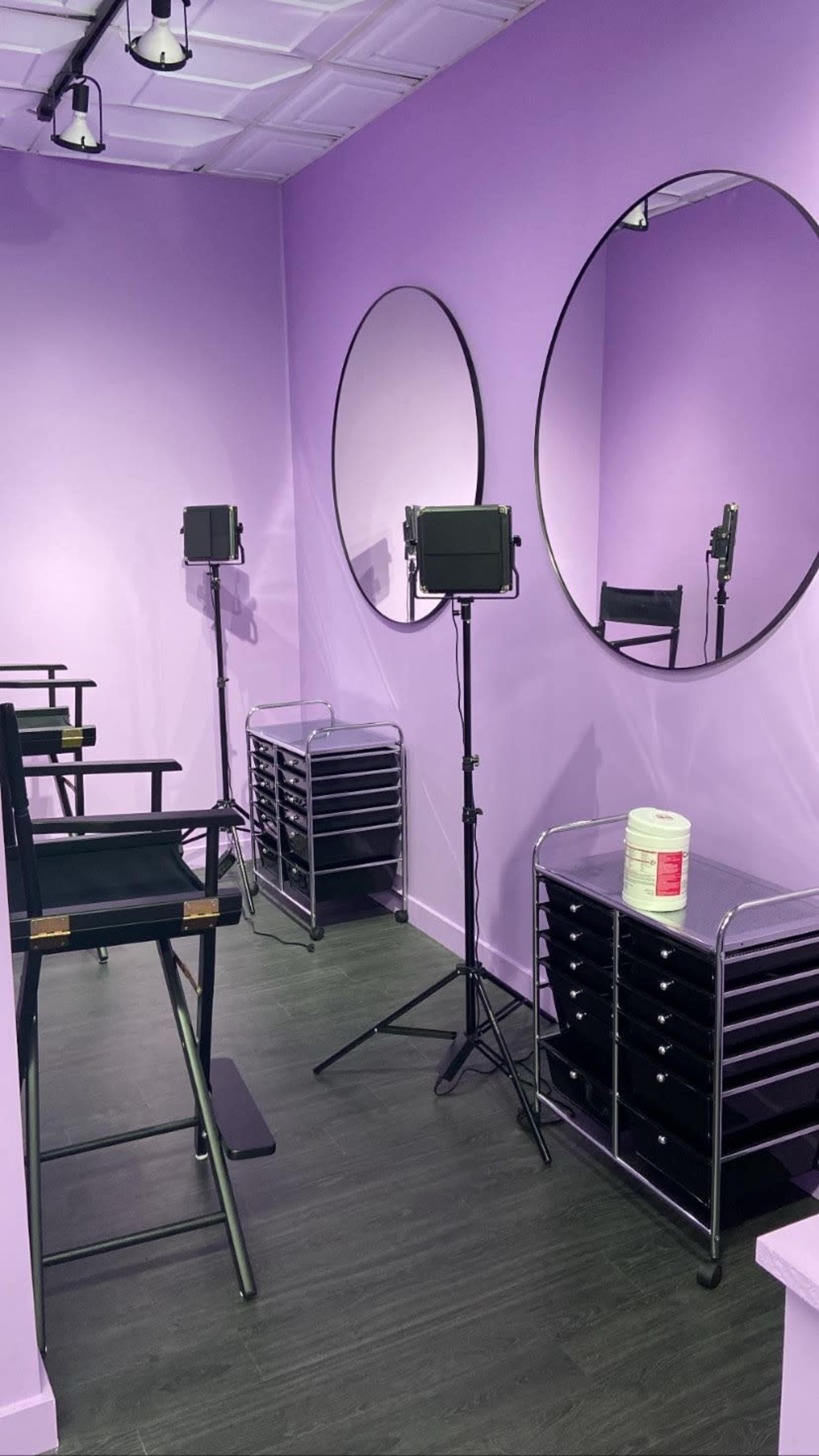 The image shows a bright purple room with two round mirrors, a pair of black director's chairs, a lighting setup on a tripod, and a storage unit with multiple drawers.