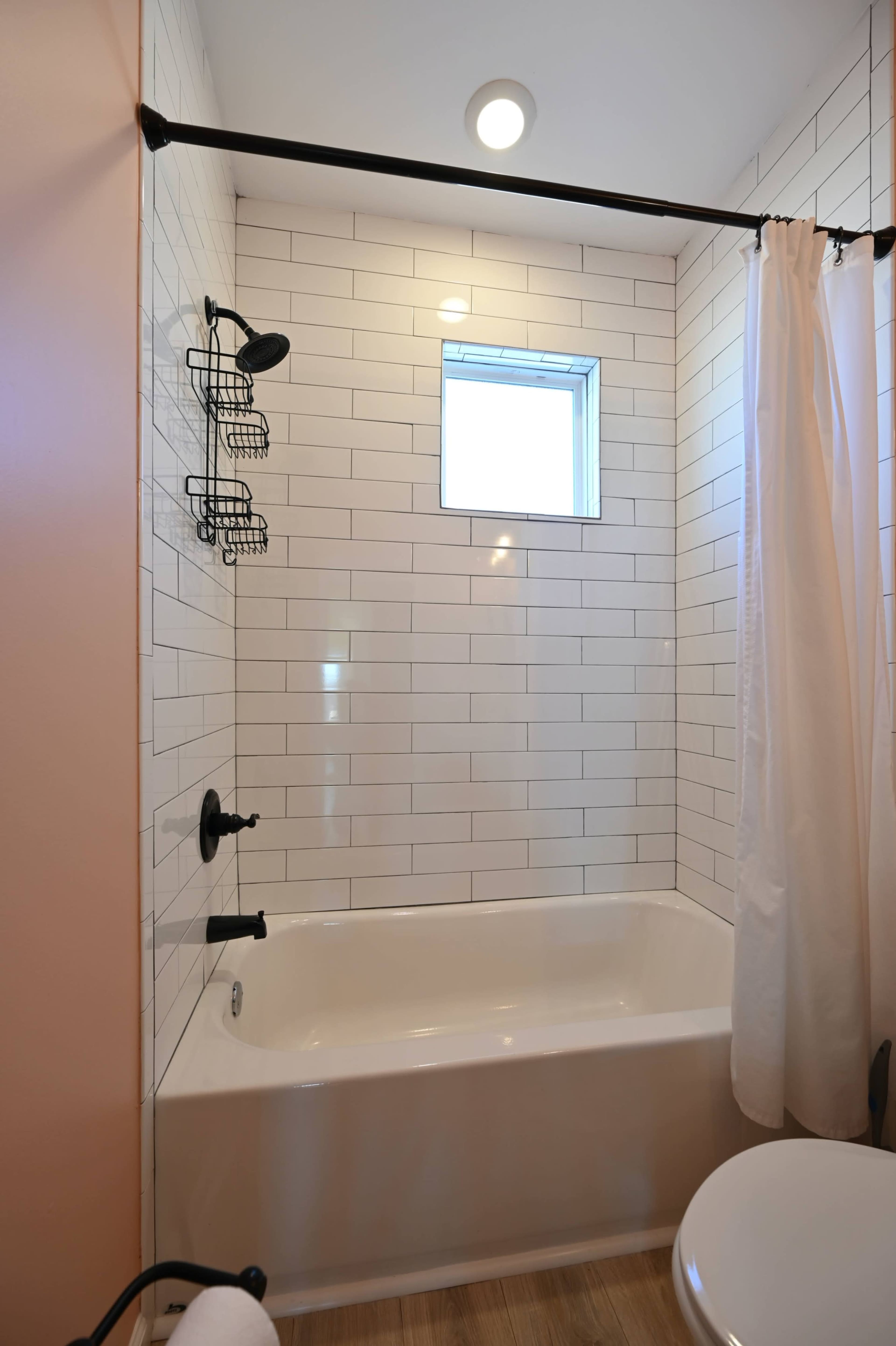 The image shows a bathroom with a white tiled shower area, a bathtub, a showerhead, and a small window.