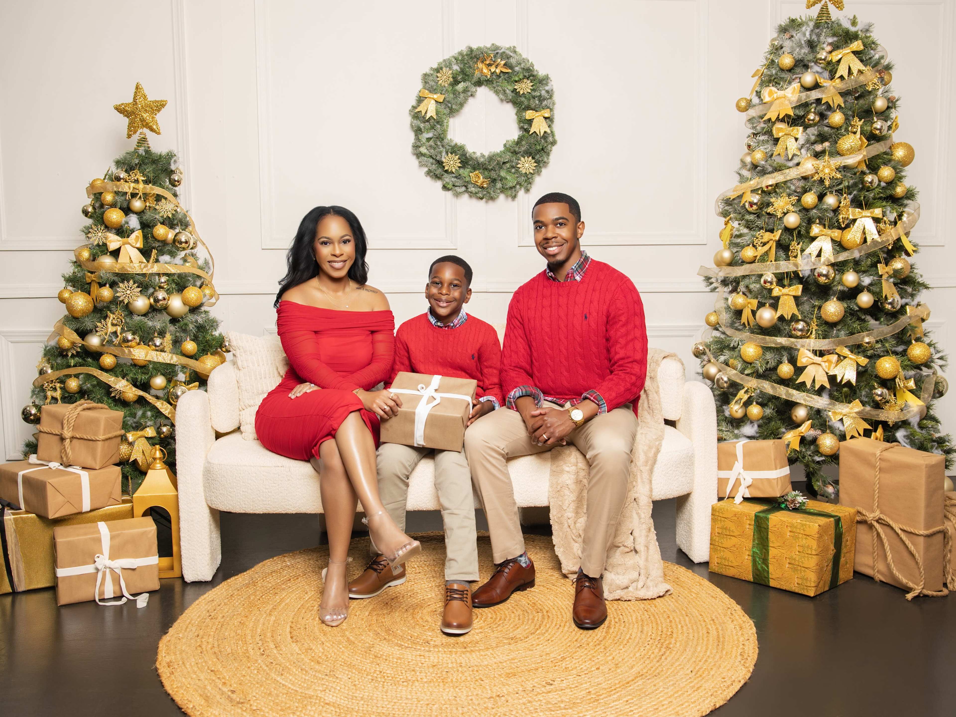 A family of three poses together on a sofa surrounded by festive decorations, including Christmas trees, a wreath, and wrapped gifts.