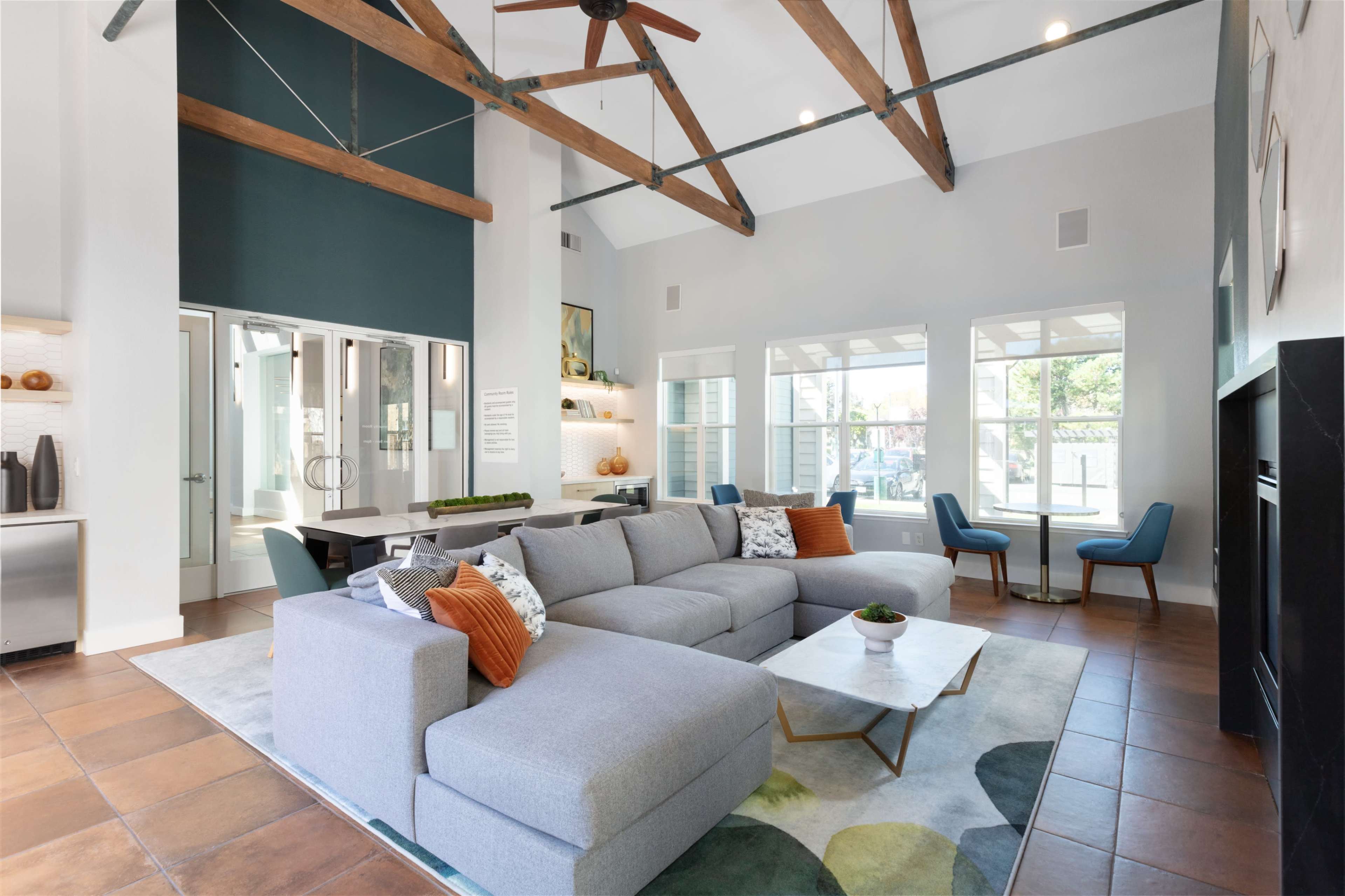 The image shows a modern living room with a large gray sectional sofa, a coffee table, and a high ceiling with exposed wooden beams.