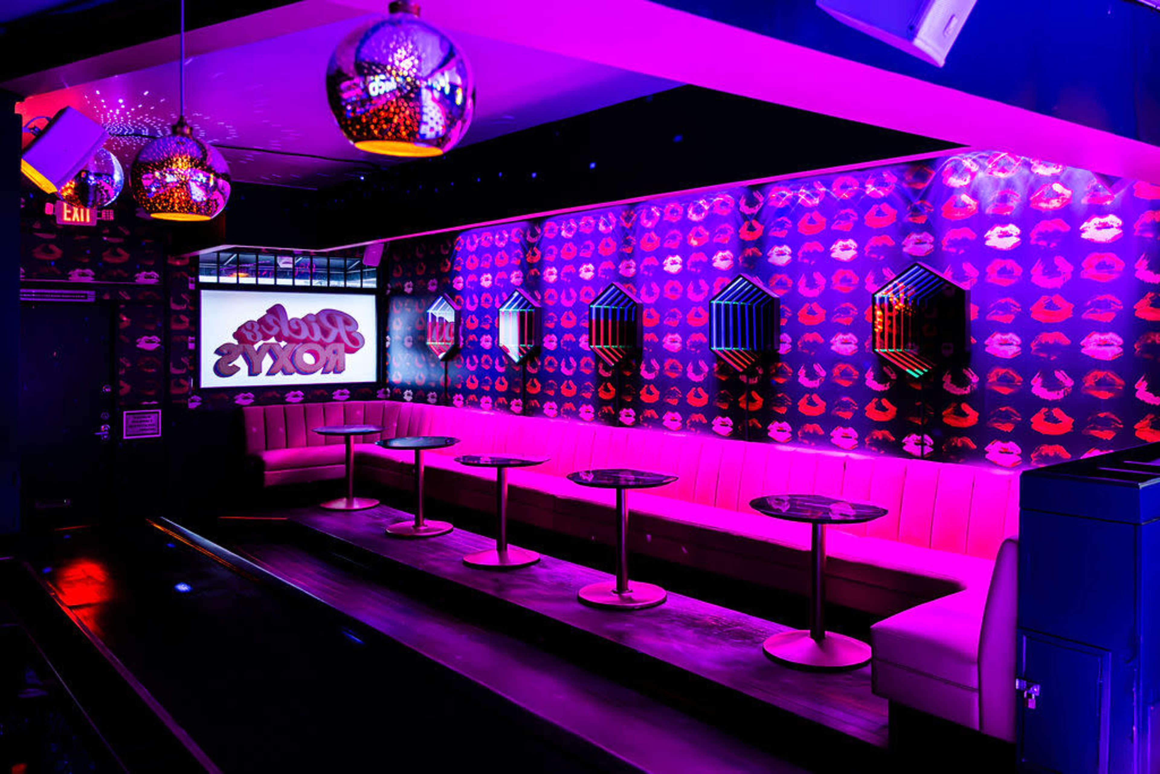The image shows a vibrant nightclub interior with pink lighting, a patterned wall covered in lips, and modern seating arranged around small tables.