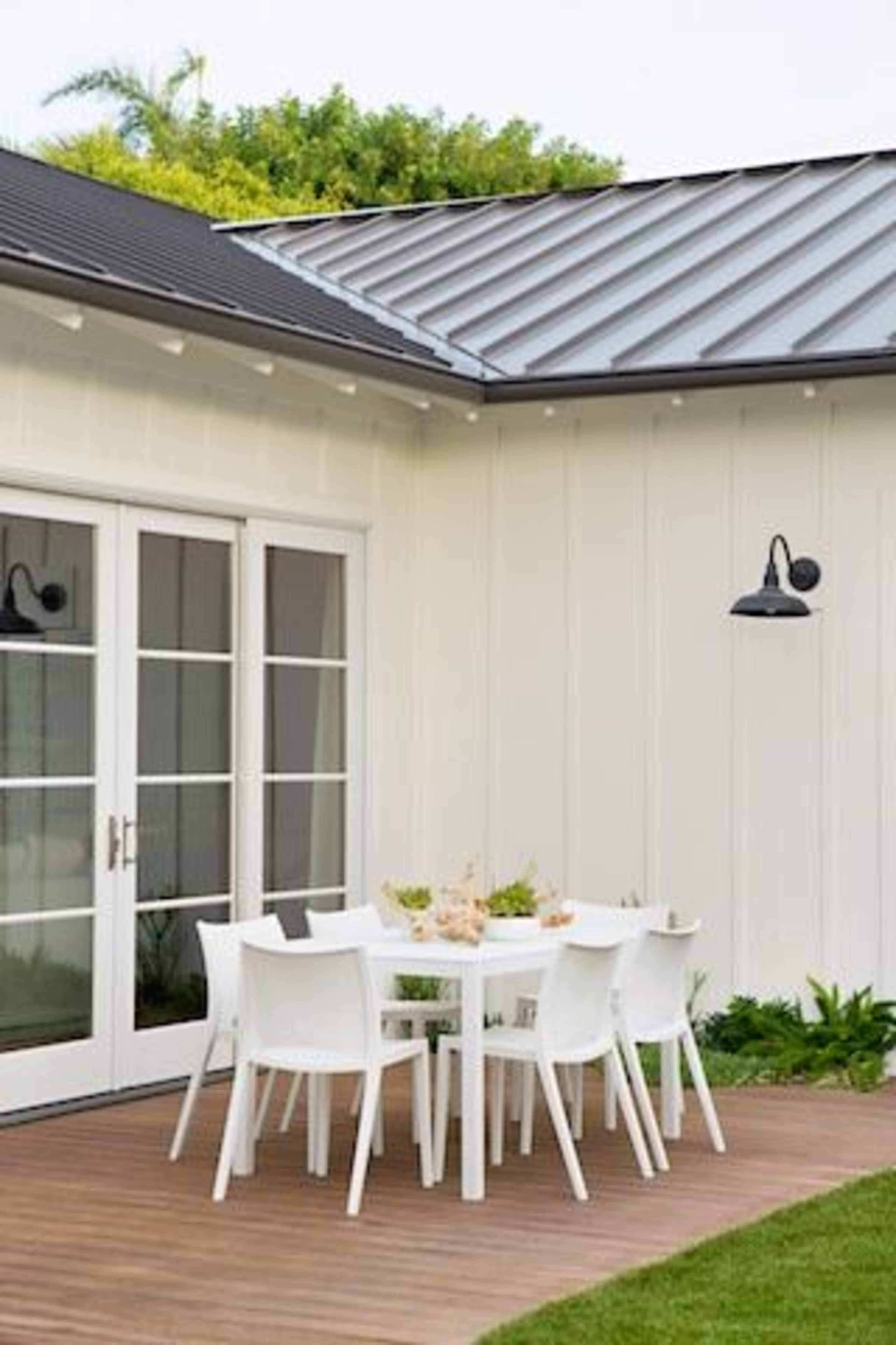A modern outdoor dining area features a white table surrounded by white chairs on a wooden deck near a light-colored wall.