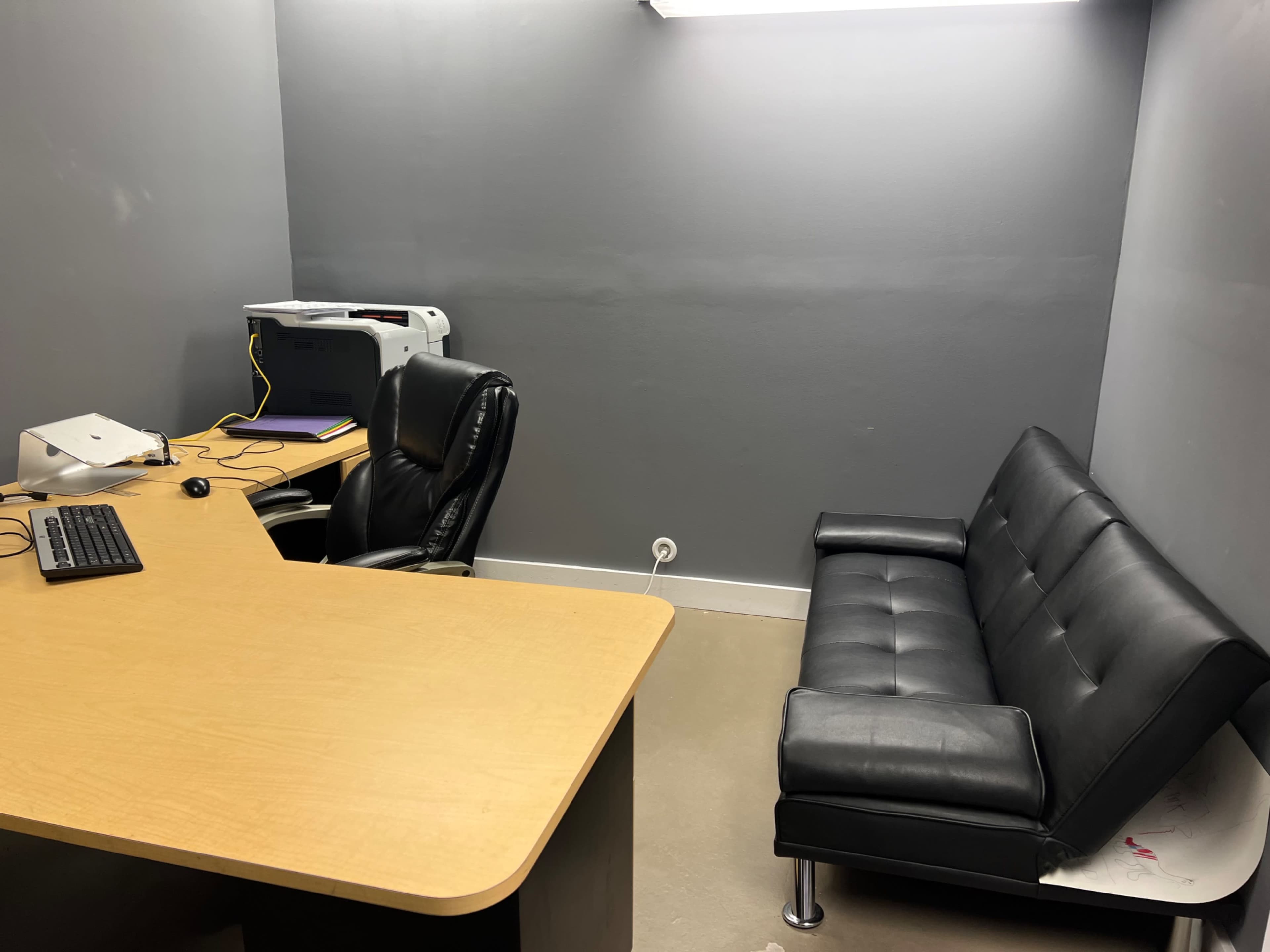 A small office with a wooden desk, an office chair, a black leather sofa, and a printer against a gray wall.