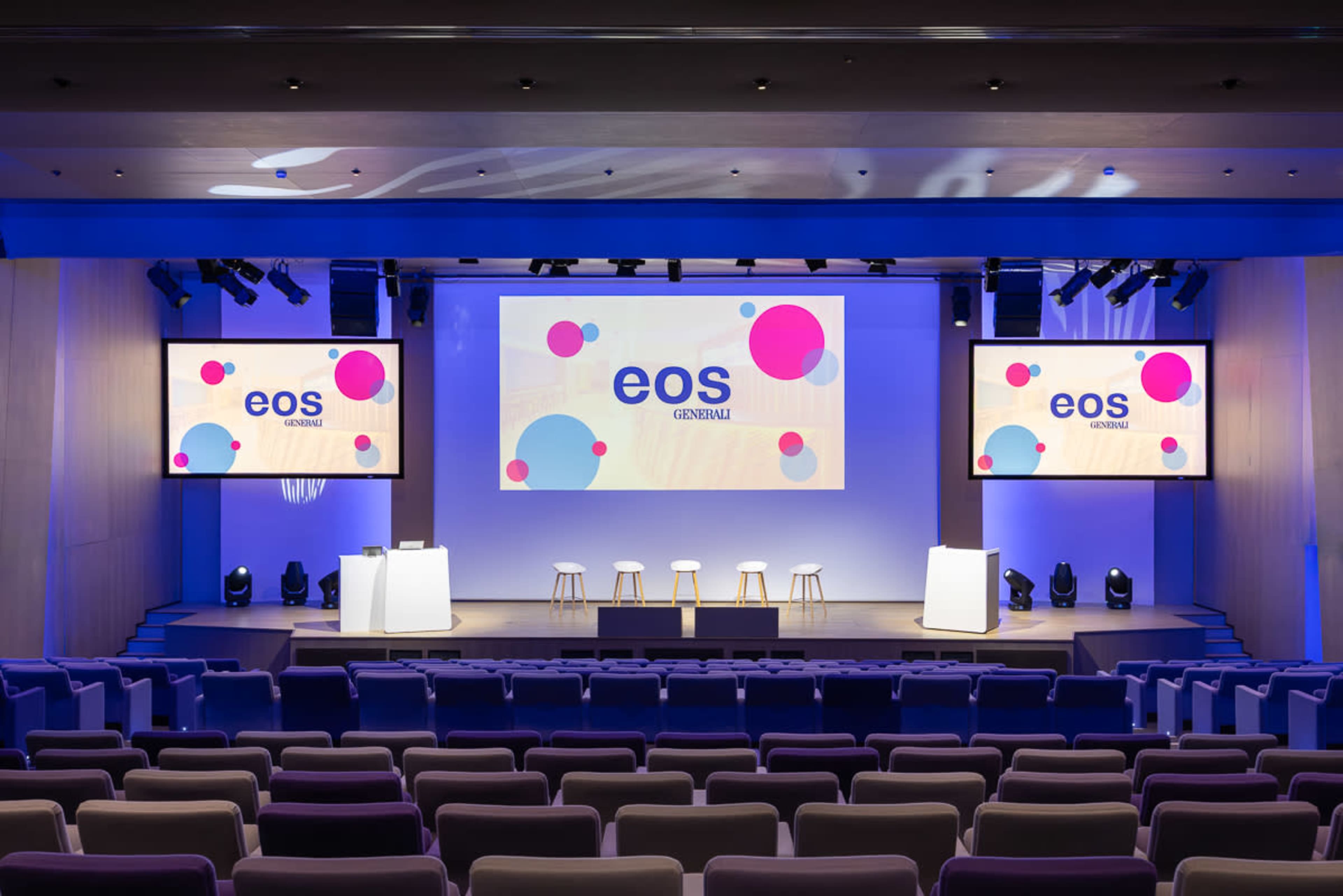 The image shows a modern conference stage with large screens displaying the logo "eos generali" and several empty seats arranged in front of it.