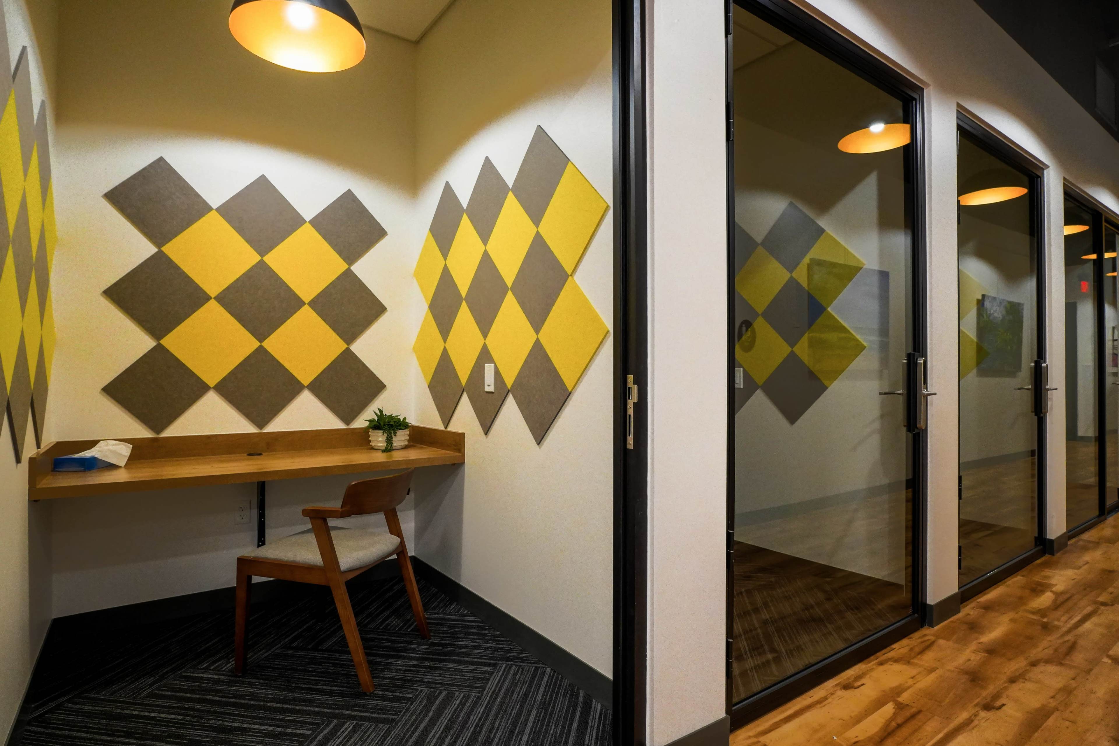A small workspace featuring a wooden desk and a chair is set against a wall adorned with yellow and gray acoustic panels, with glass-enclosed offices visible in the background.