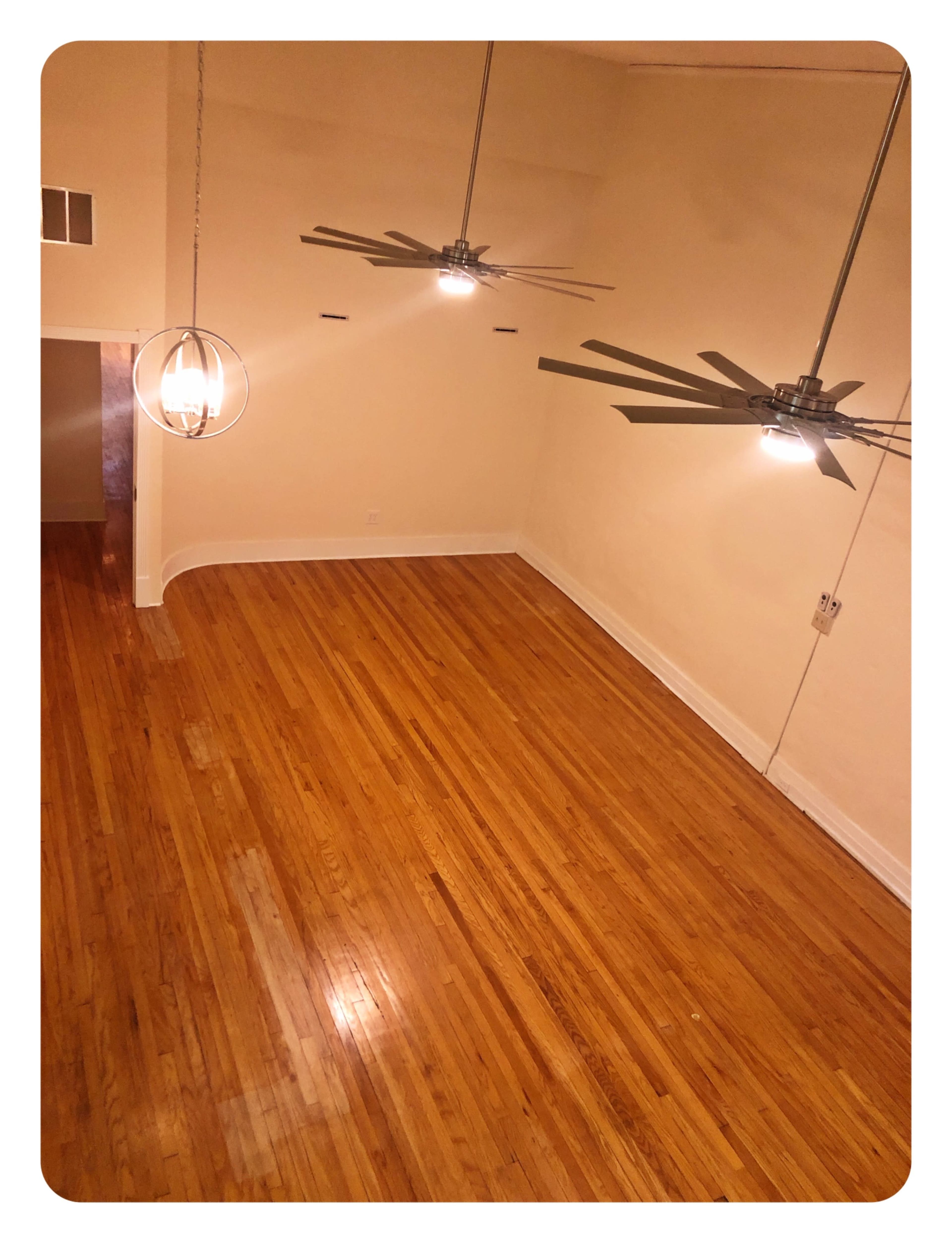 A spacious room with hardwood flooring, two ceiling fans, and warm lighting.