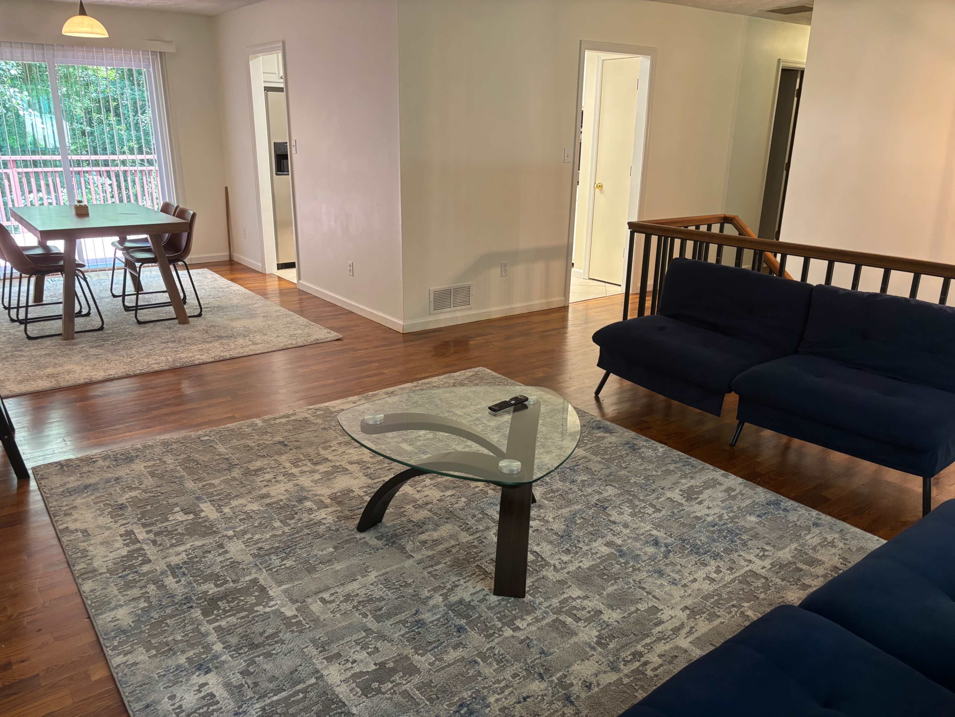 The image shows a living room with a dark blue couch and a glass coffee table on a patterned area rug, adjacent to a dining area with a wooden table and chairs.