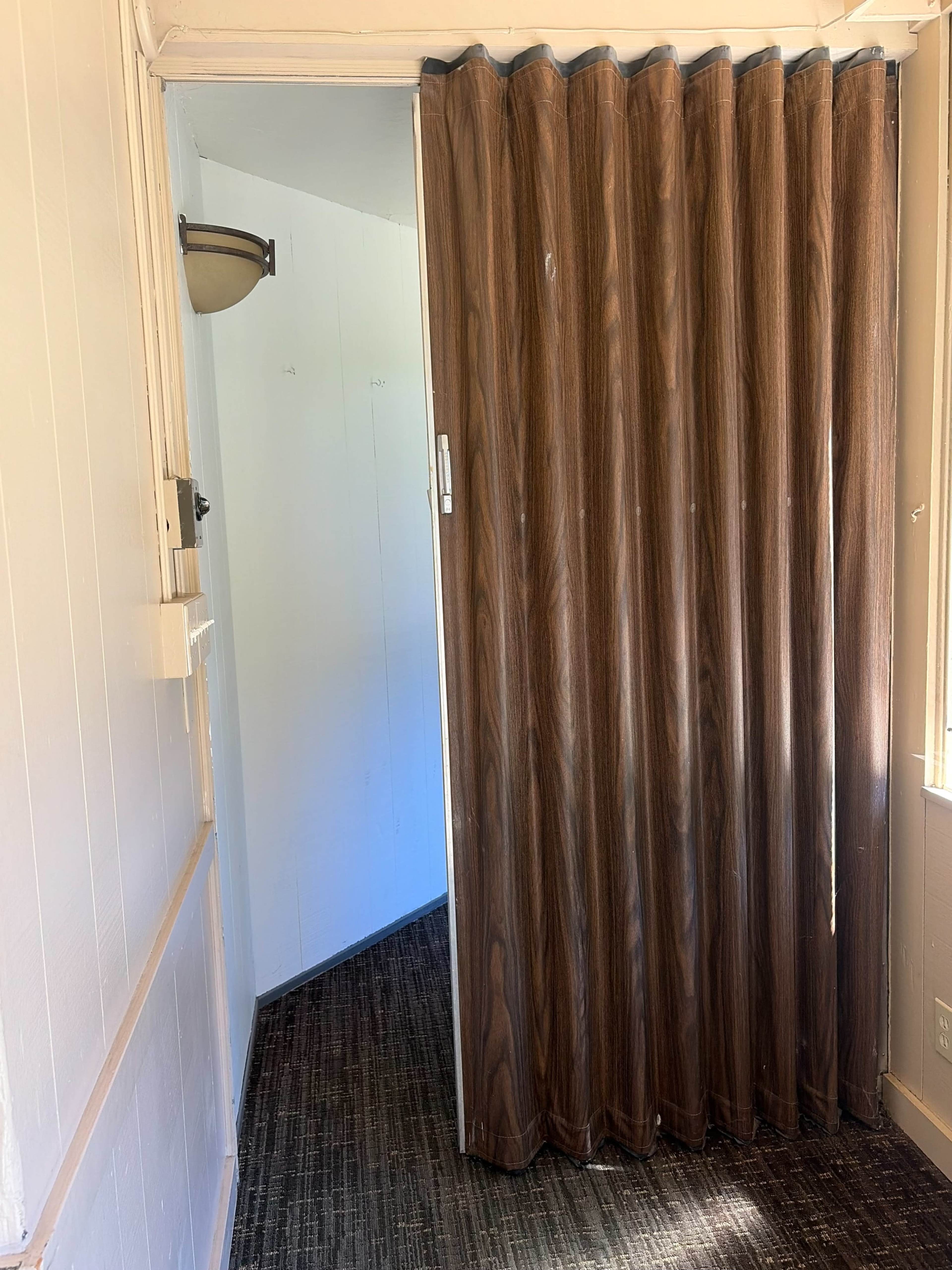 The image shows a doorway with a brown accordion-style curtain partially open, leading to a lighter-colored room.