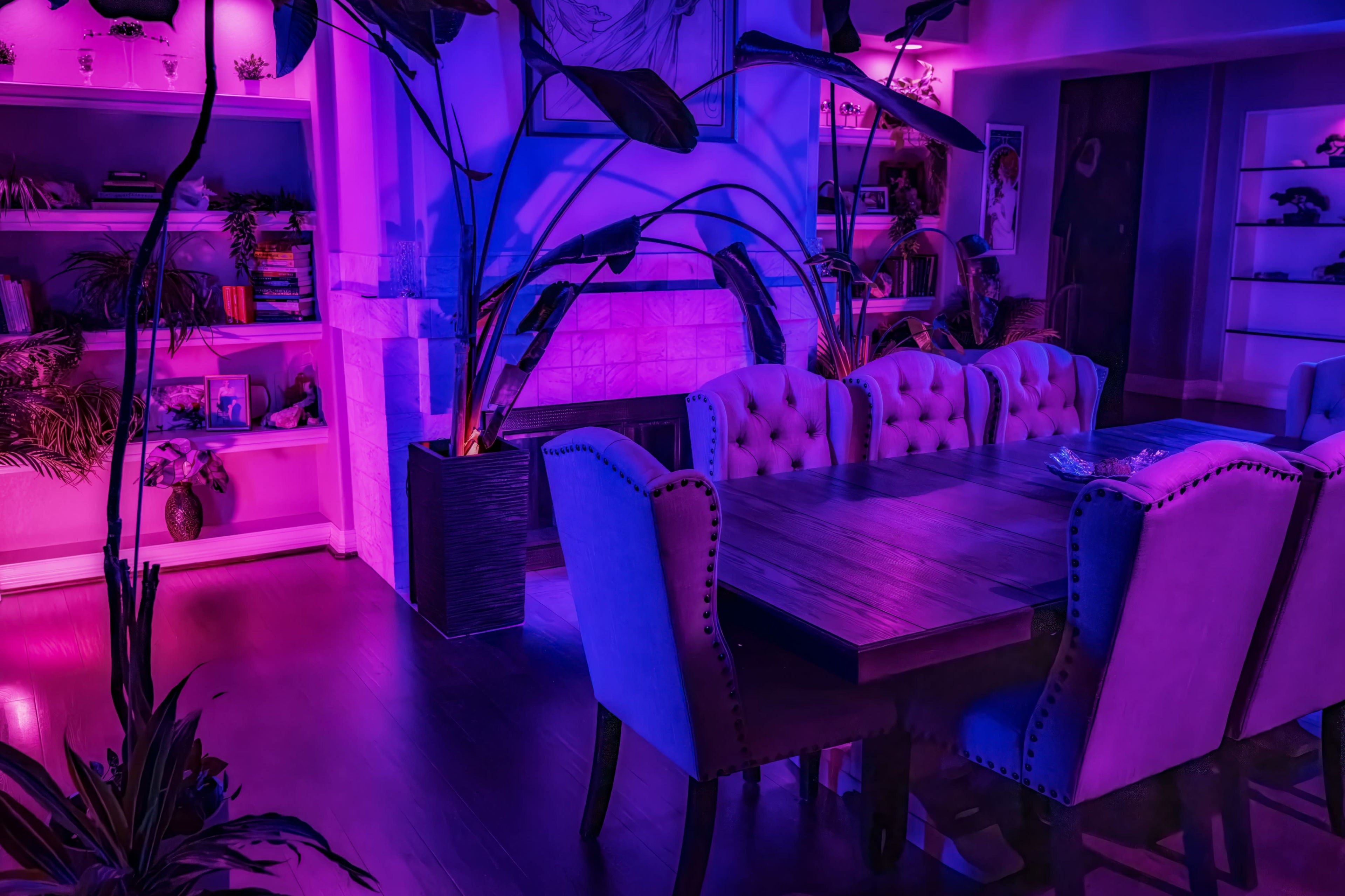 A dining room with a wooden table and upholstered chairs, illuminated by purple and blue lights, features a fireplace and shelves adorned with plants and decorative items.