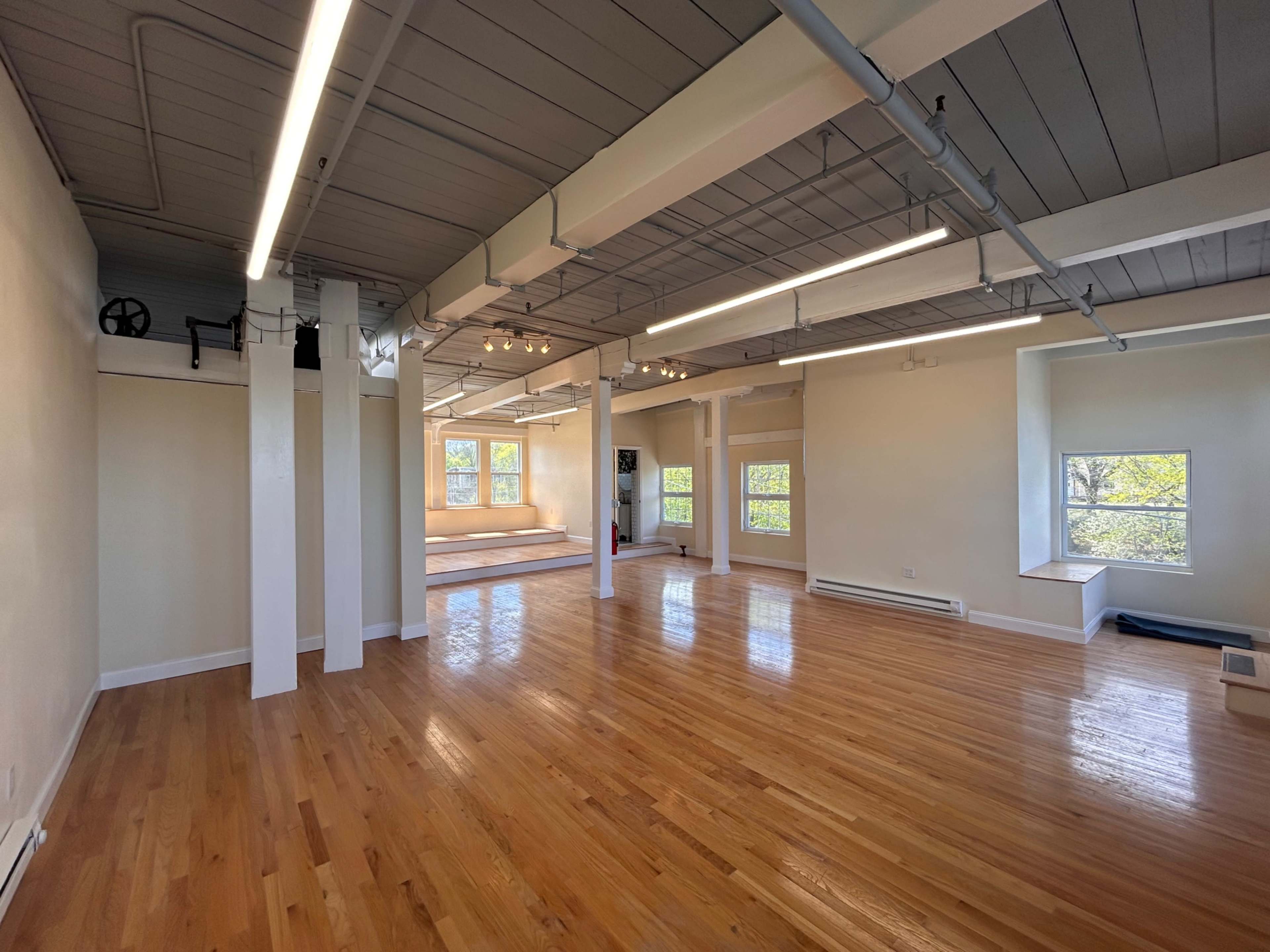 The image shows a spacious, well-lit room with hardwood floors, large windows, and minimal furnishings.