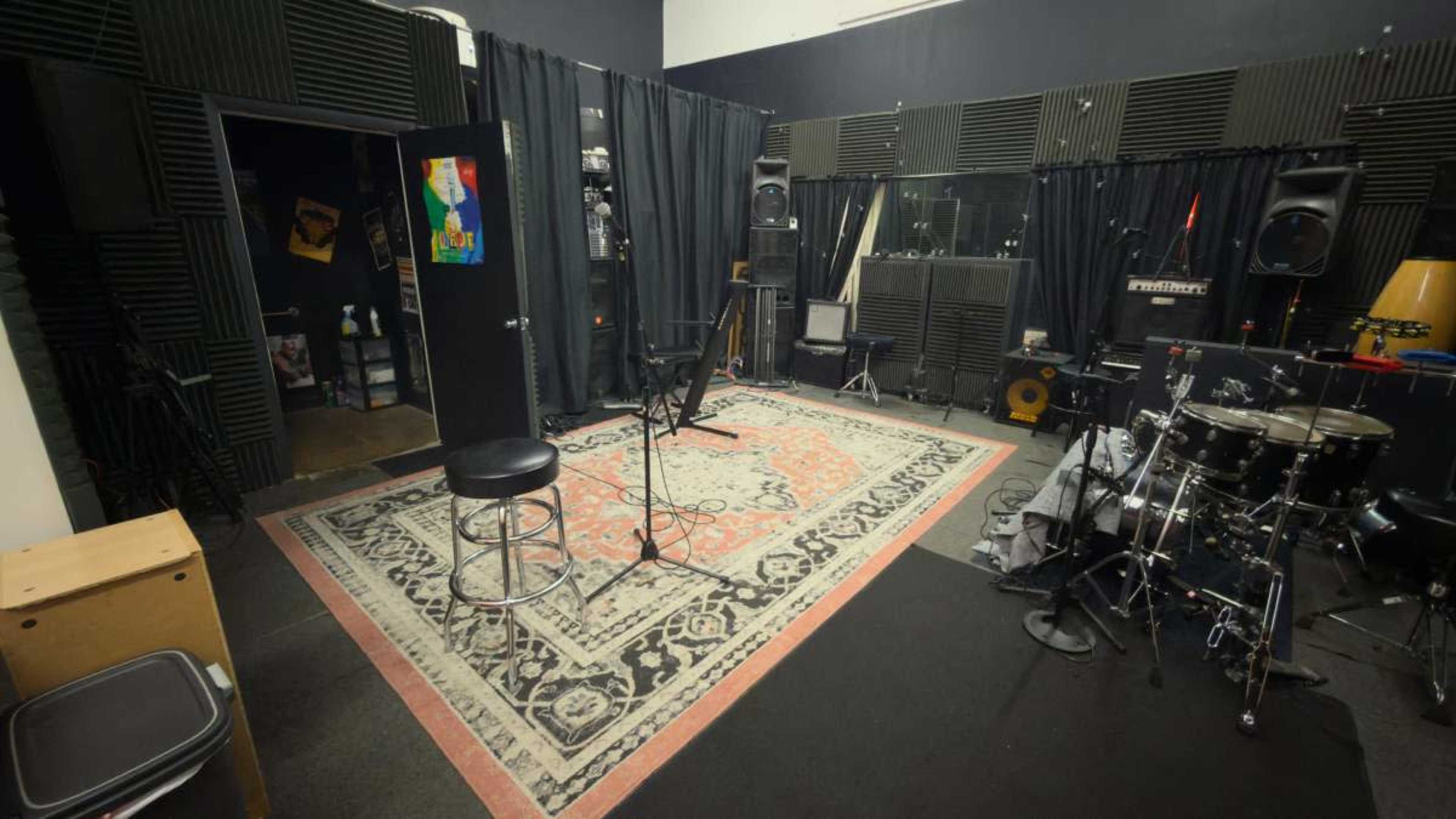 The image shows a recording studio with a drum set, microphone, and soundproofing panels, along with a patterned rug and a stool.