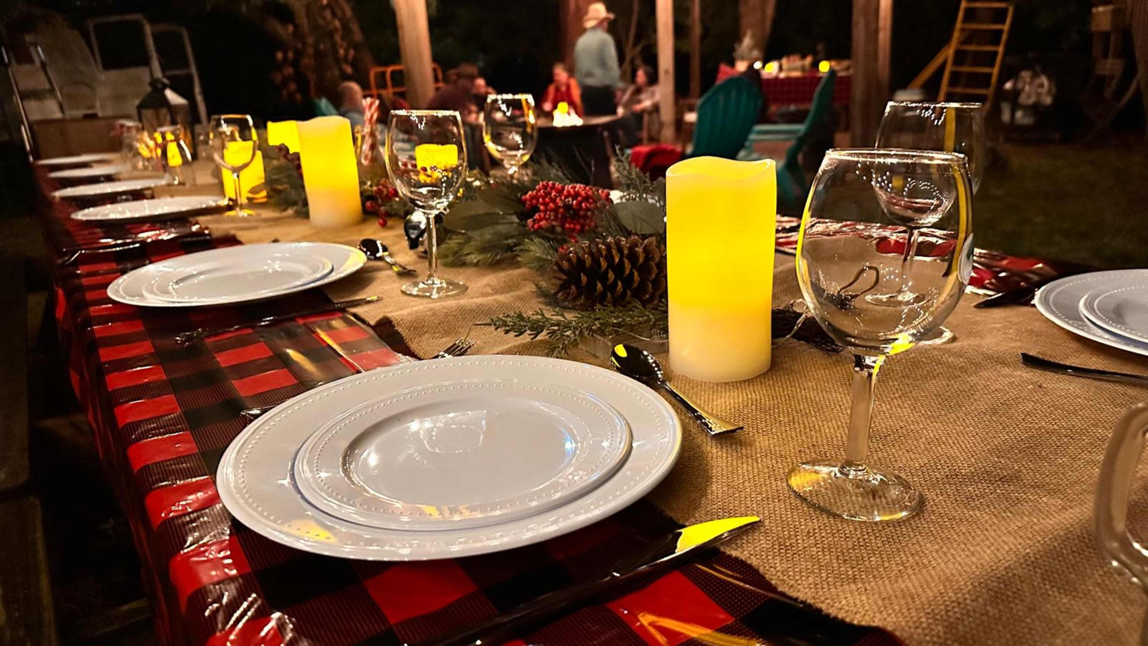 A long dining table is set outdoors with white plates, glasses, and a lit candle centerpiece, surrounded by guests in a festive atmosphere.