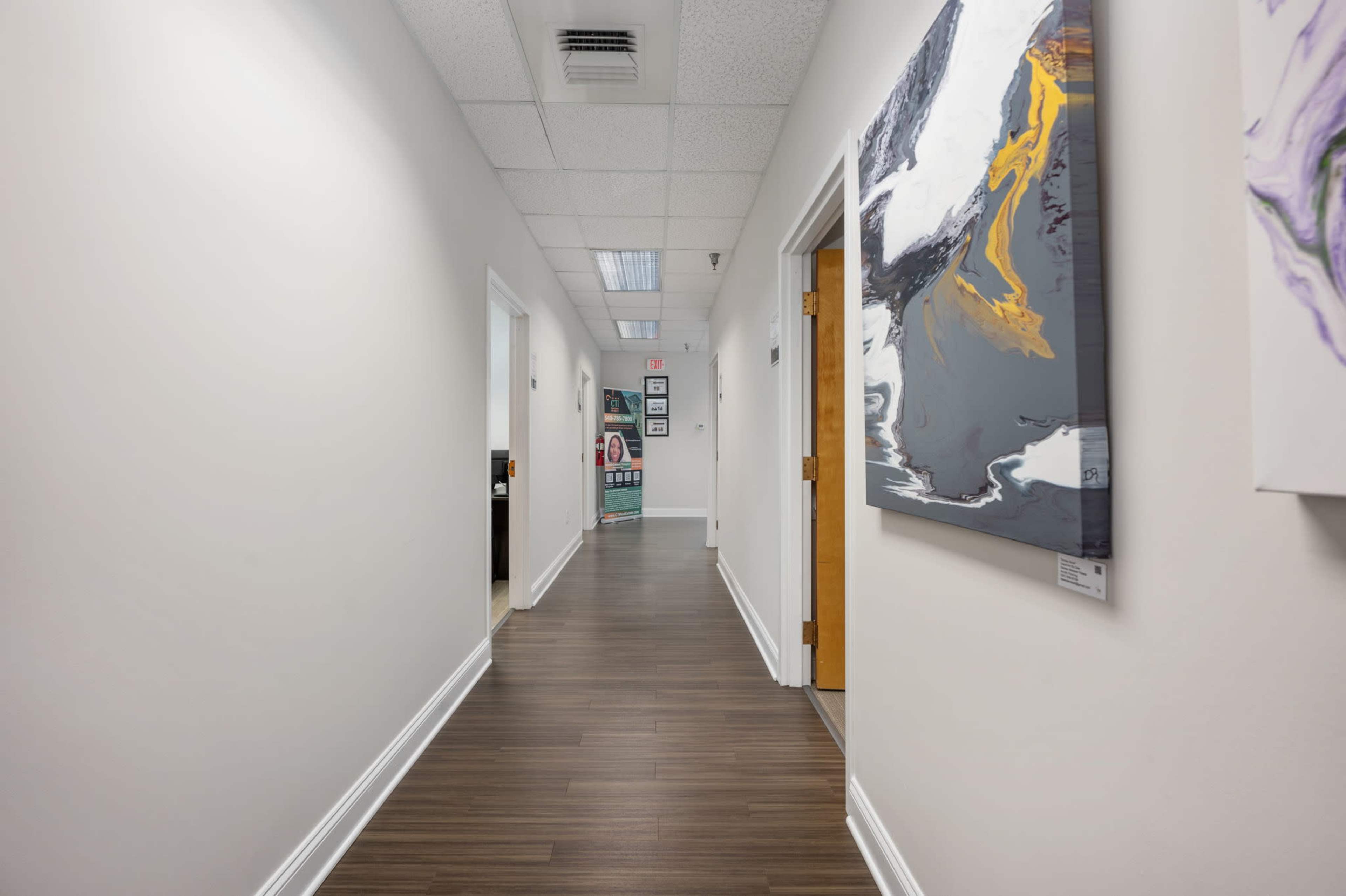 A hallway with wooden flooring, white walls, and doorways on either side, leading to an office space at the end.