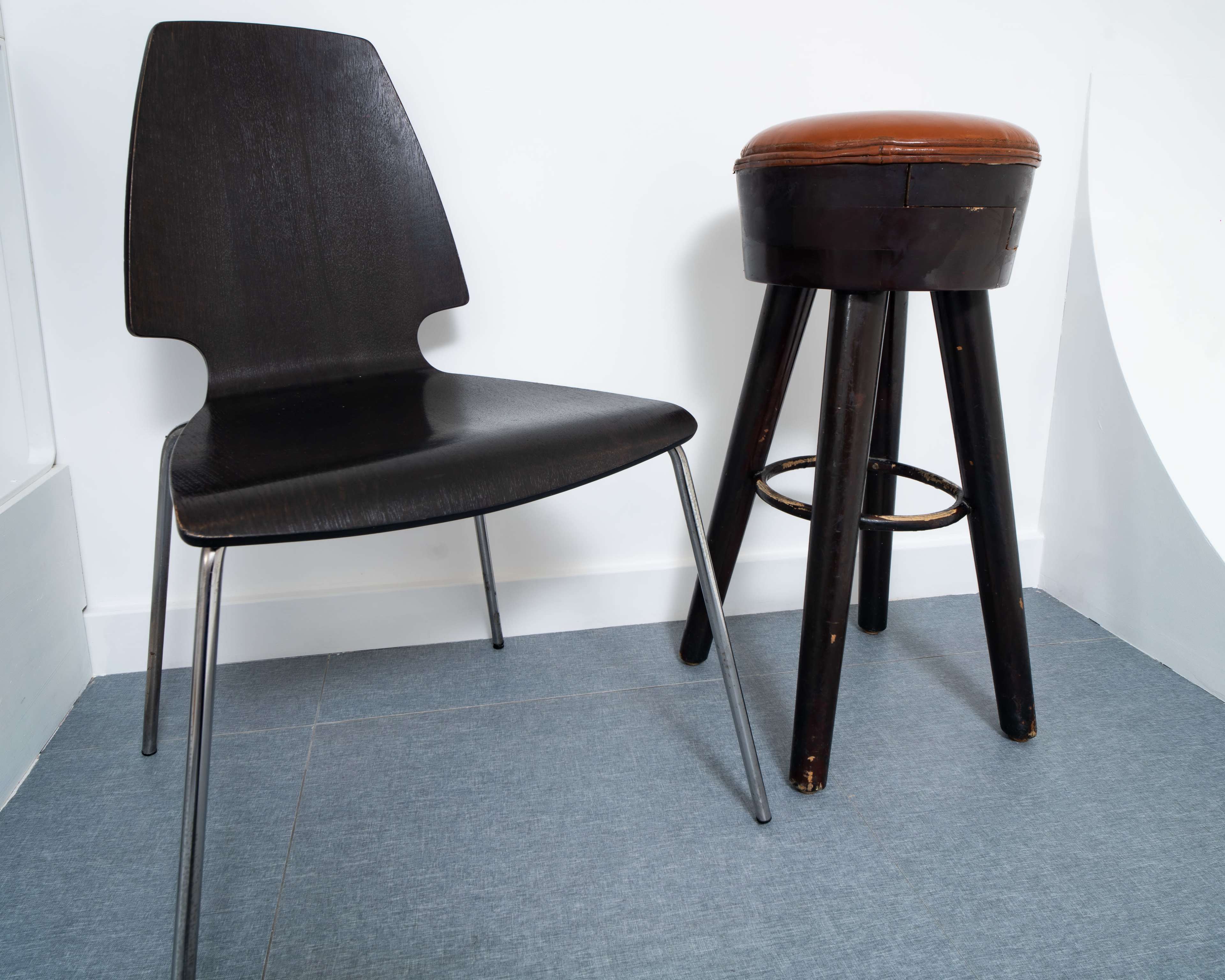 A black chair and a wooden stool with a brown cushion are placed in a corner on a light blue floor.
