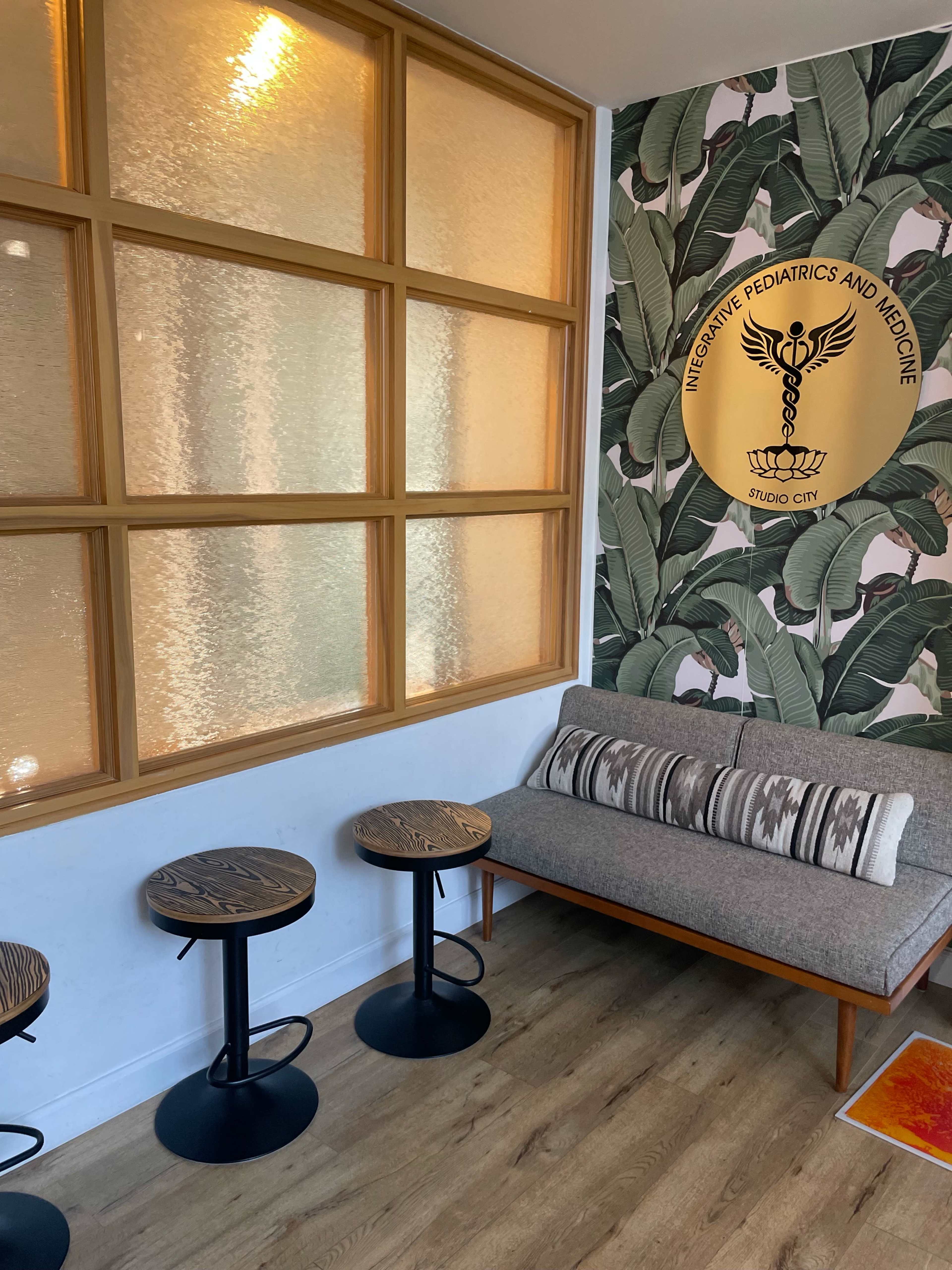 A waiting area with a gray sofa, three small round tables, a wall with a tropical leaf pattern, and a circular emblem for "Integrative Pediatrics and Medicine" on the wall.