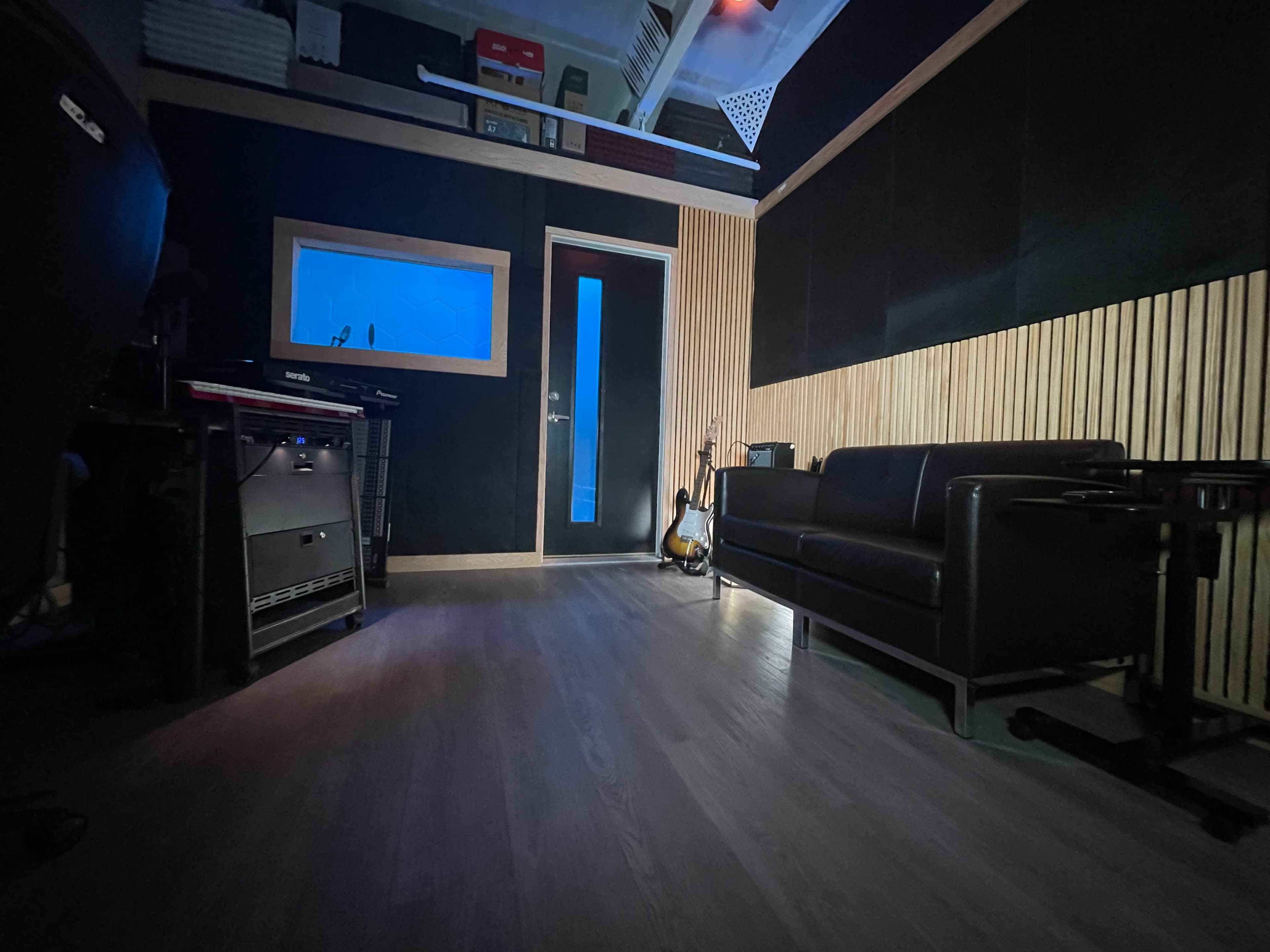 The image shows a music studio featuring a black sofa, a wooden accent wall, and a door with a blue window.