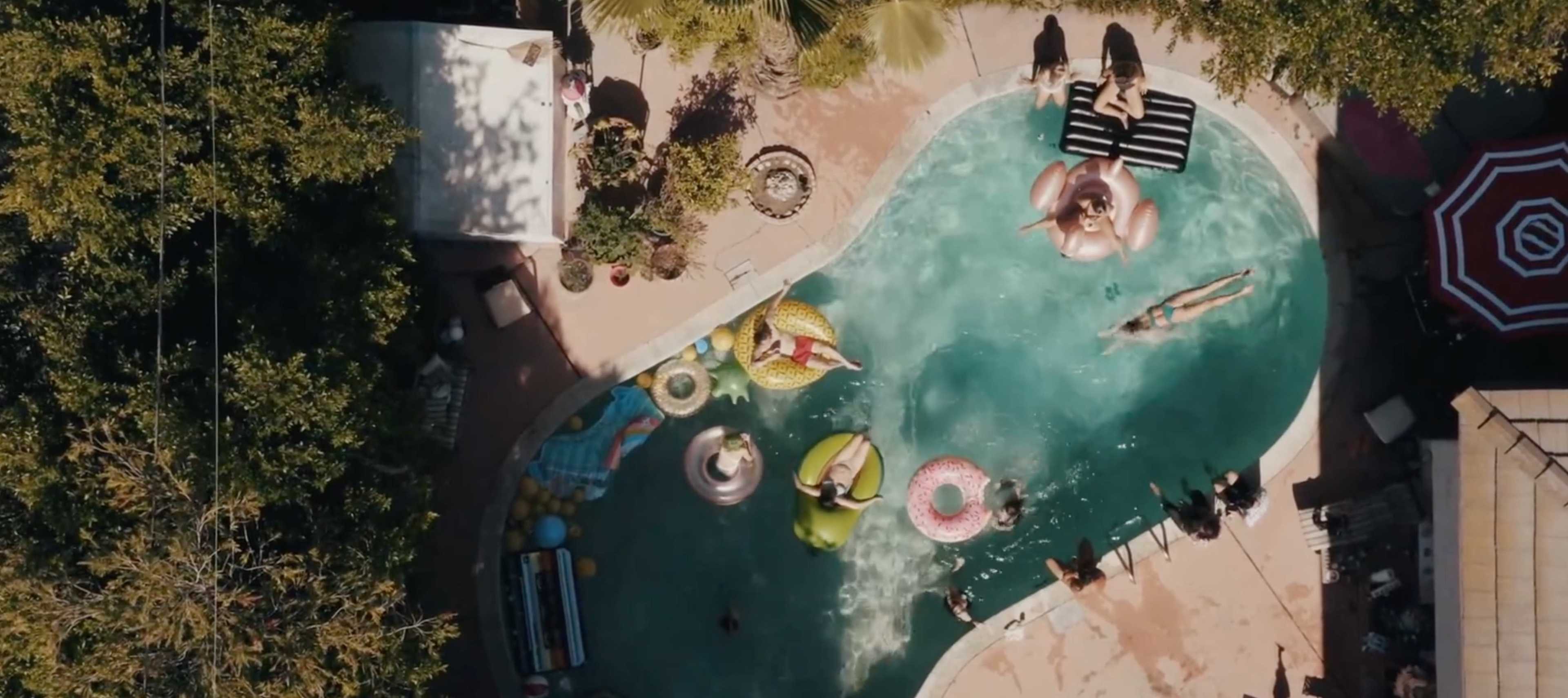 Aerial view of a swimming pool surrounded by lounge chairs, inflatables, and people enjoying leisure time.