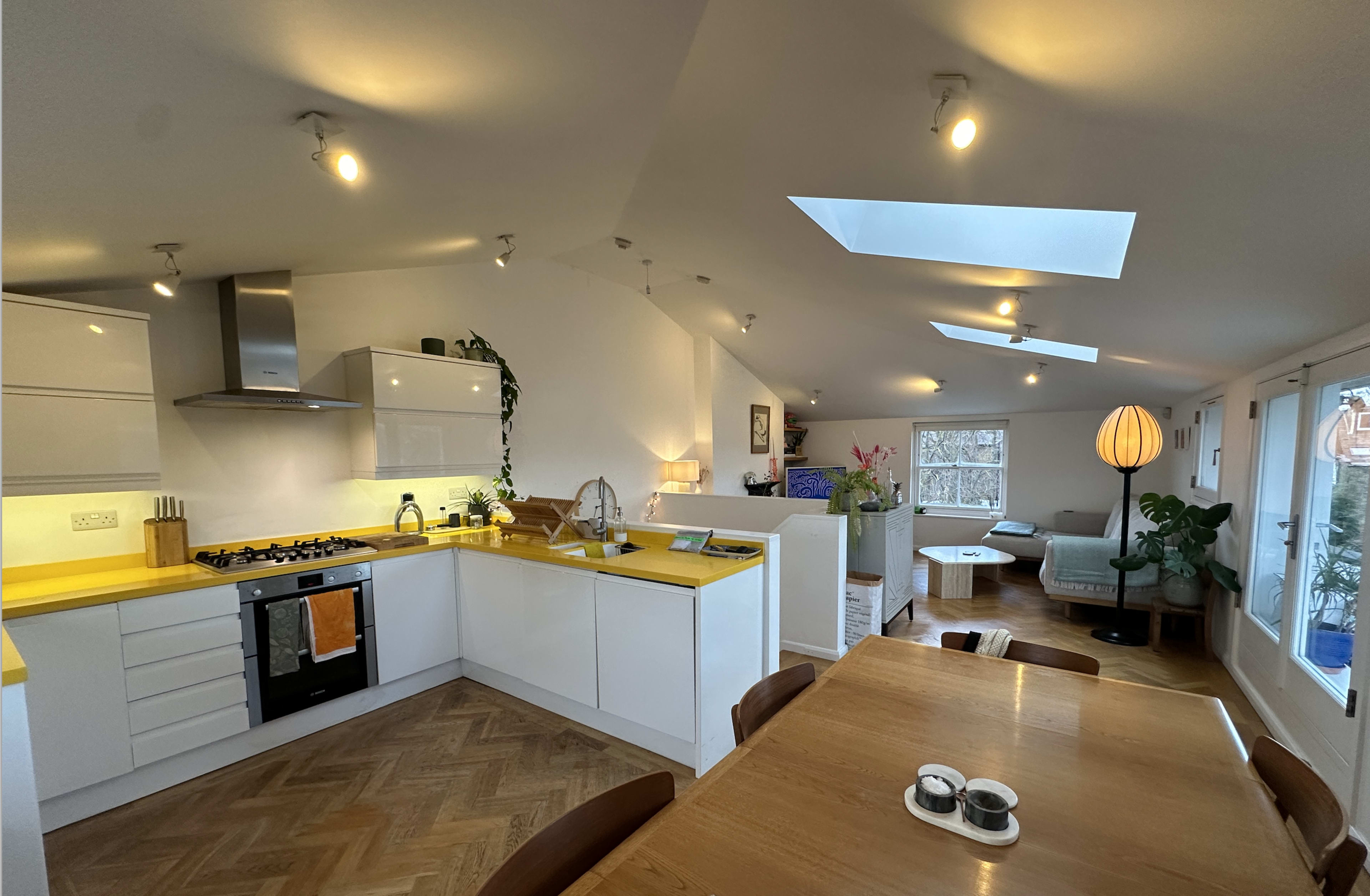 The image shows a modern kitchen with white cabinetry and yellow countertops, adjacent to a living area featuring a large window and a comfortable seating arrangement.
