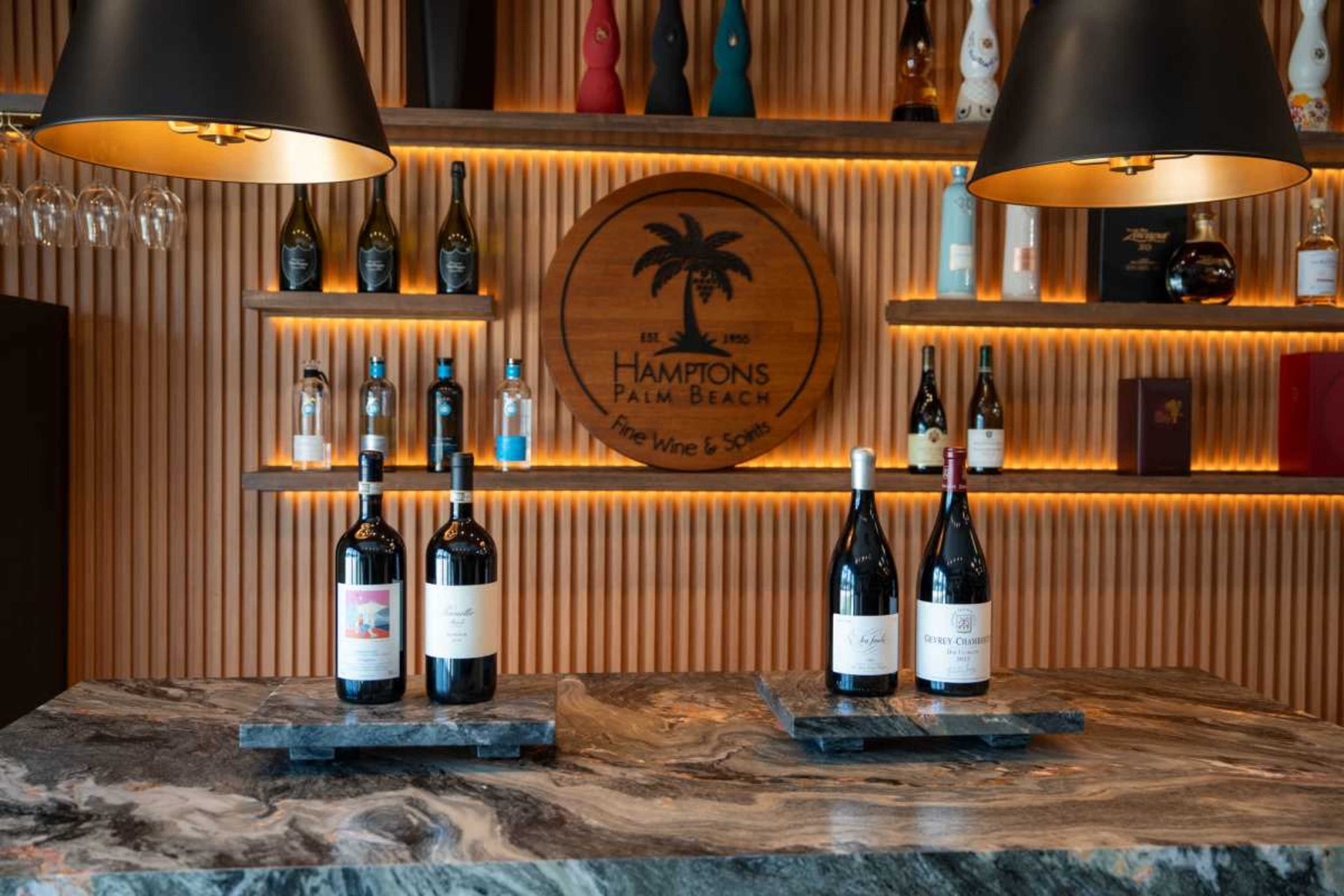 The image shows a wine display with three bottles positioned on a marble countertop in front of a wooden bar featuring various wine bottles and a circular logo reading "Hamptons Palm Beach."