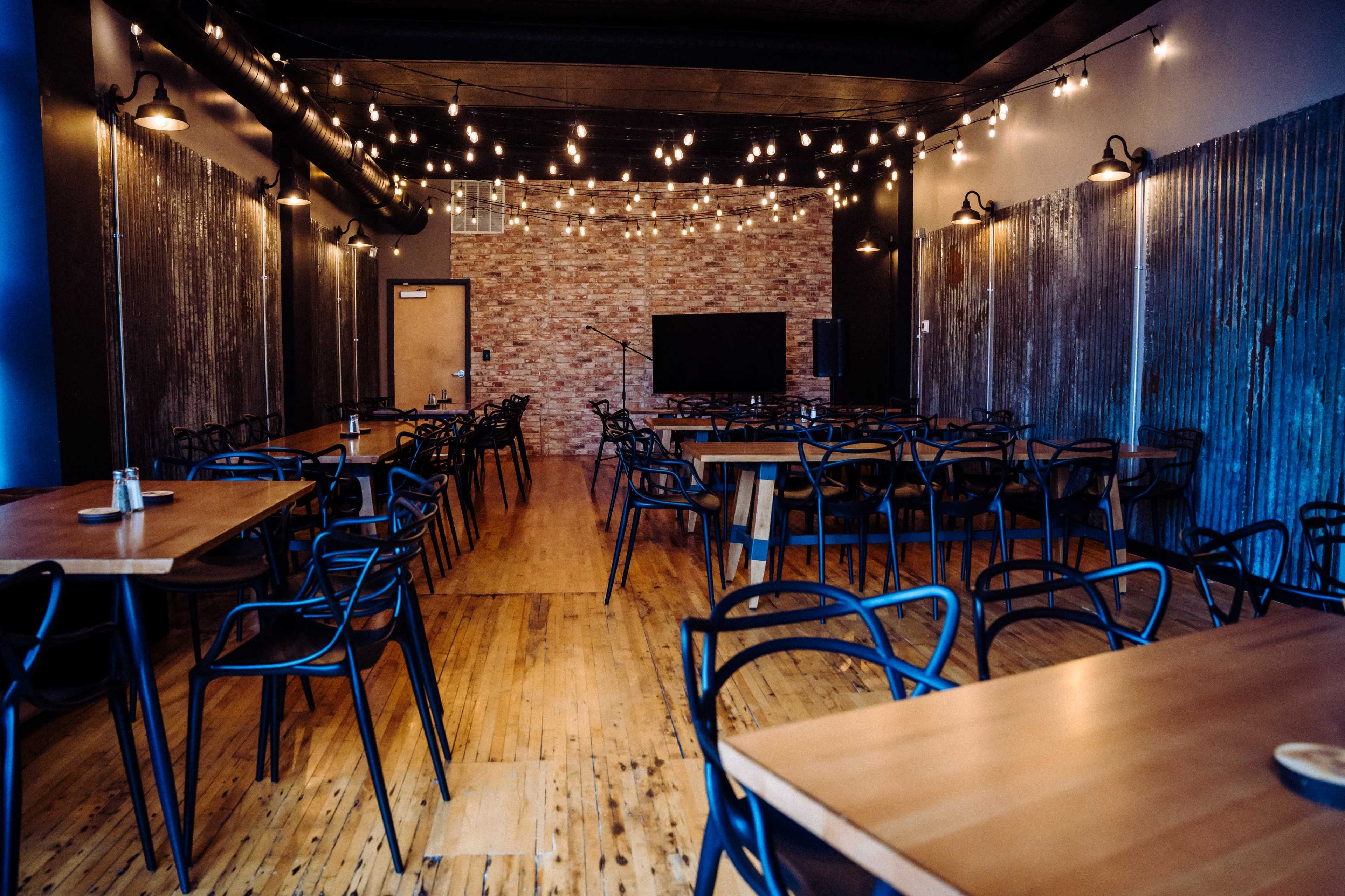 The image shows an empty restaurant interior with wooden floors, several tables, and string lights hanging from the ceiling.