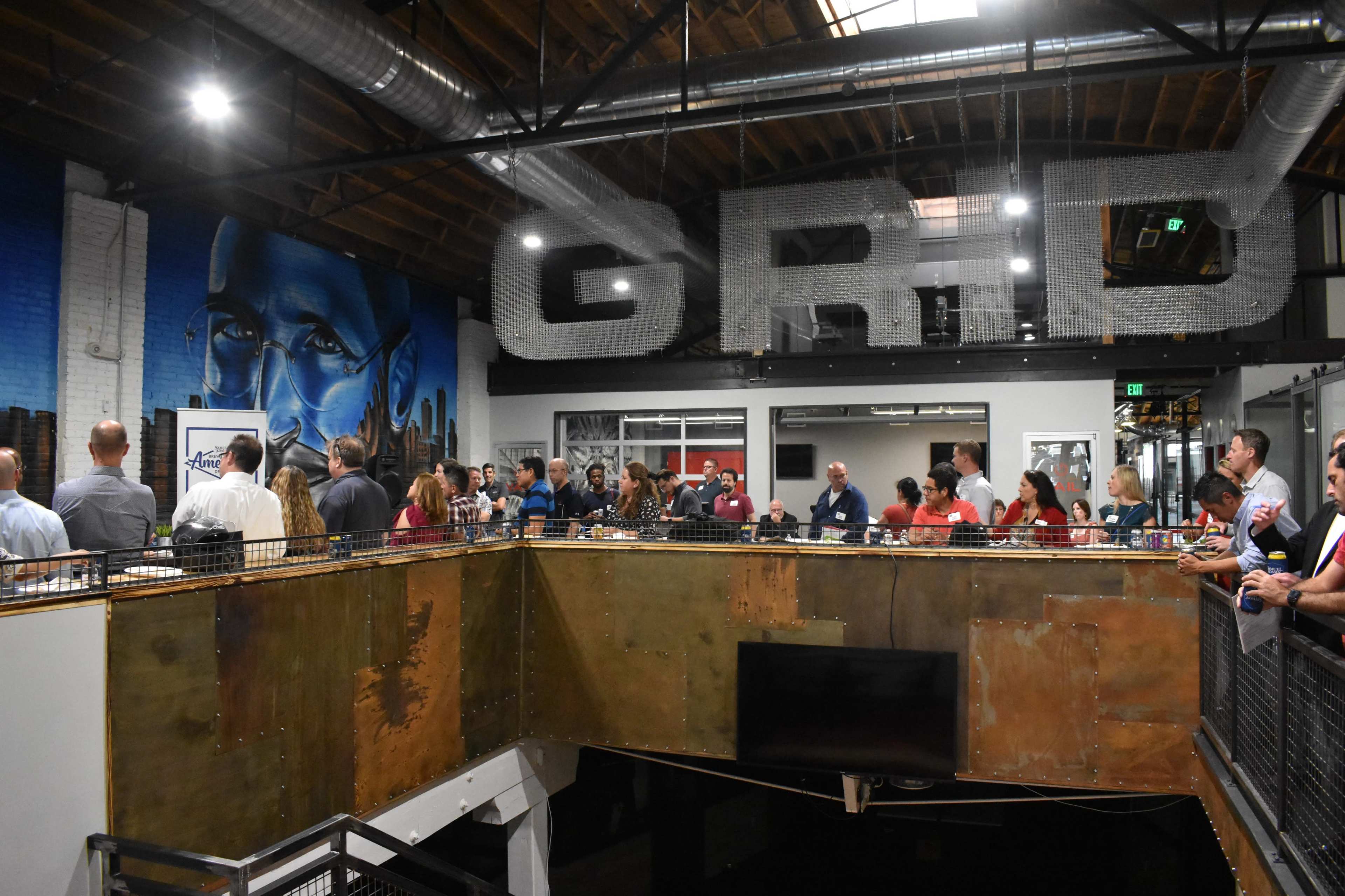 A large group of people gathers on an upper level in a modern space with industrial decor and a prominent mural on the wall.