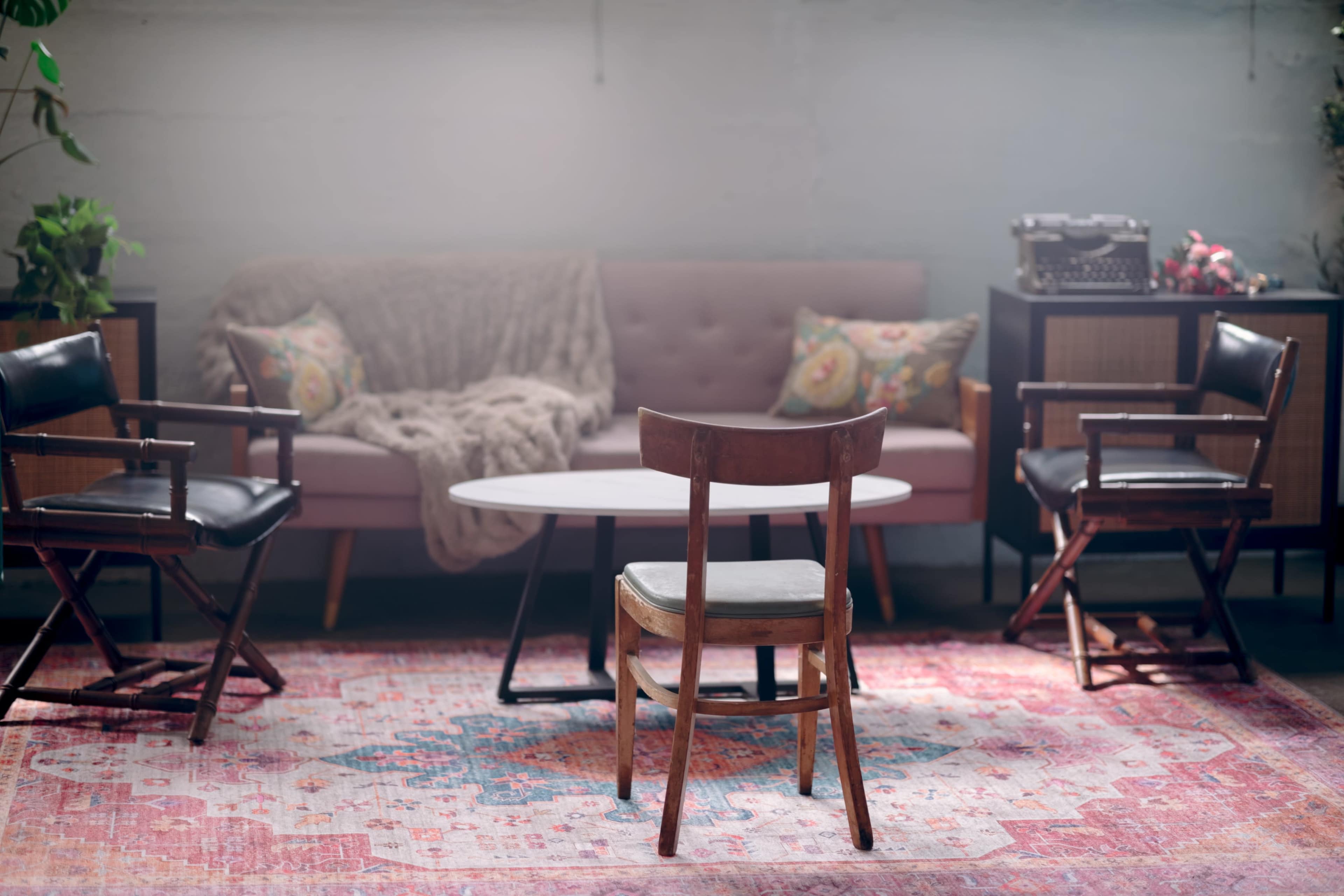 A cozy living room features a pink sofa, a round coffee table, and several wooden chairs arranged around a patterned rug.