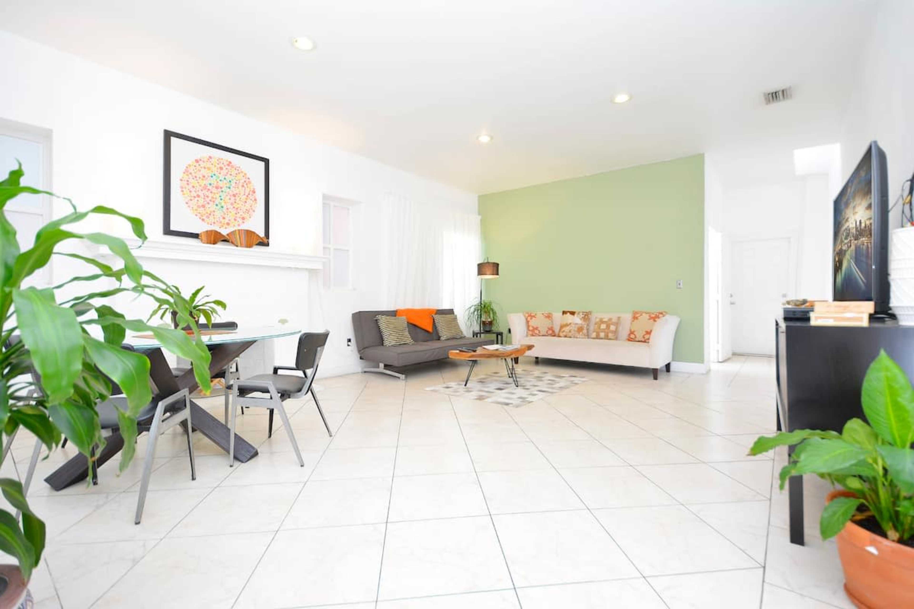 A bright, modern living space with a gray sofa, a dining table with chairs, and a green accent wall.