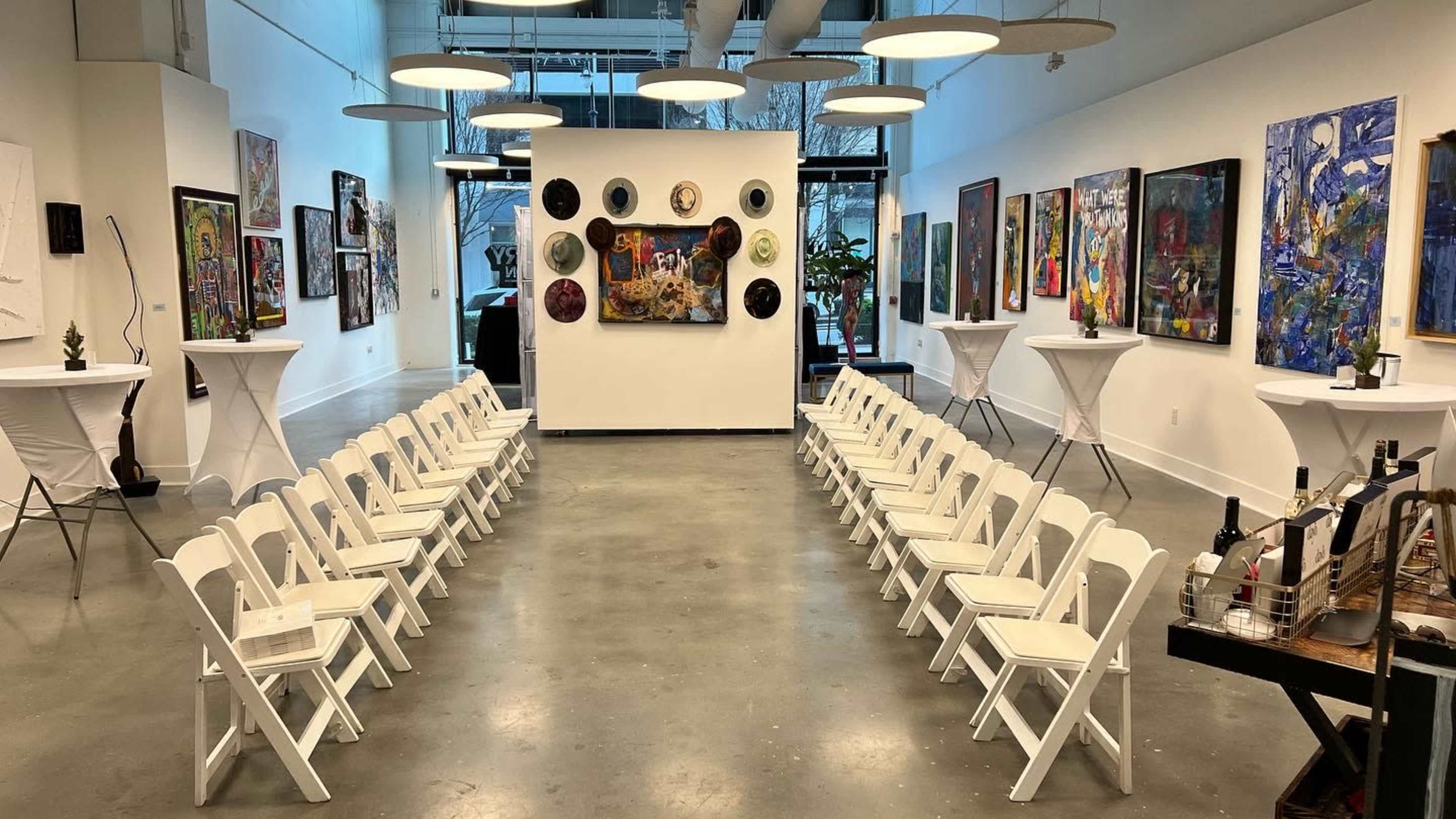 An art gallery with rows of white folding chairs facing a central wall adorned with various artworks, surrounded by additional pieces on the side walls.