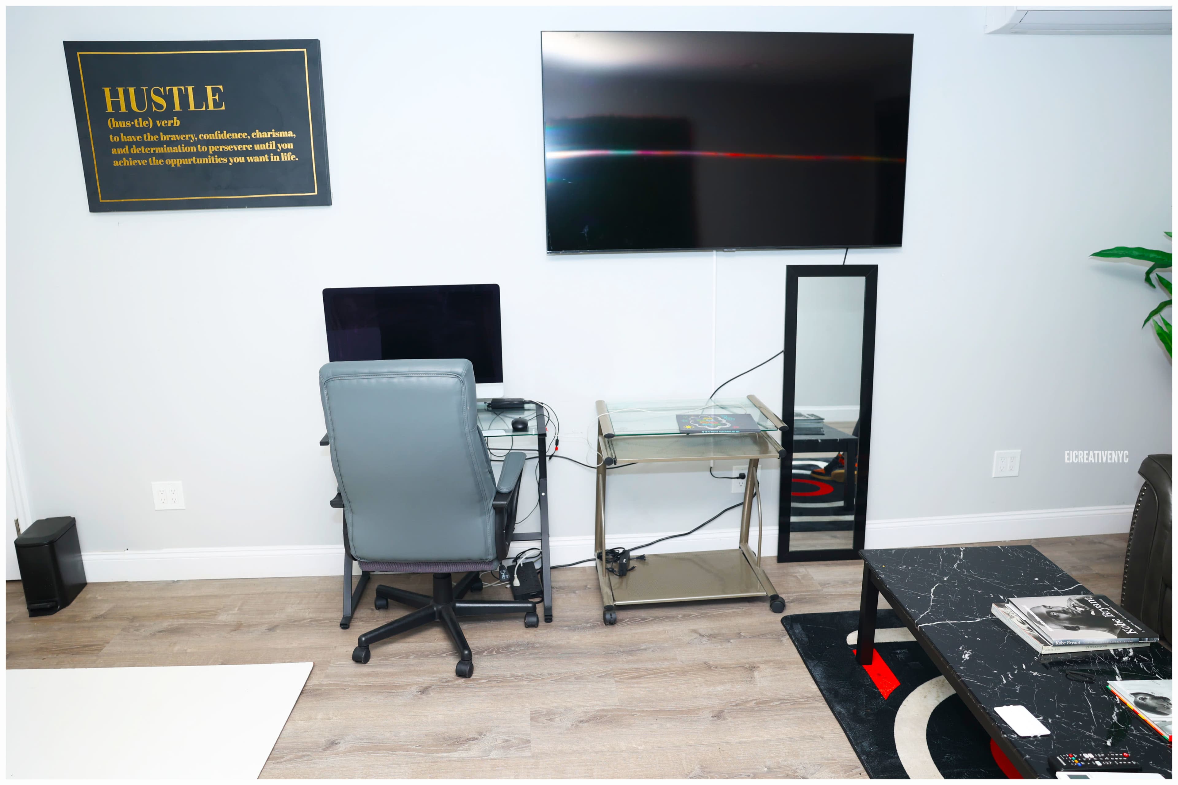 A workspace featuring a computer desk with a chair, a large wall-mounted TV, a mirror, and a coffee table with magazines on it.