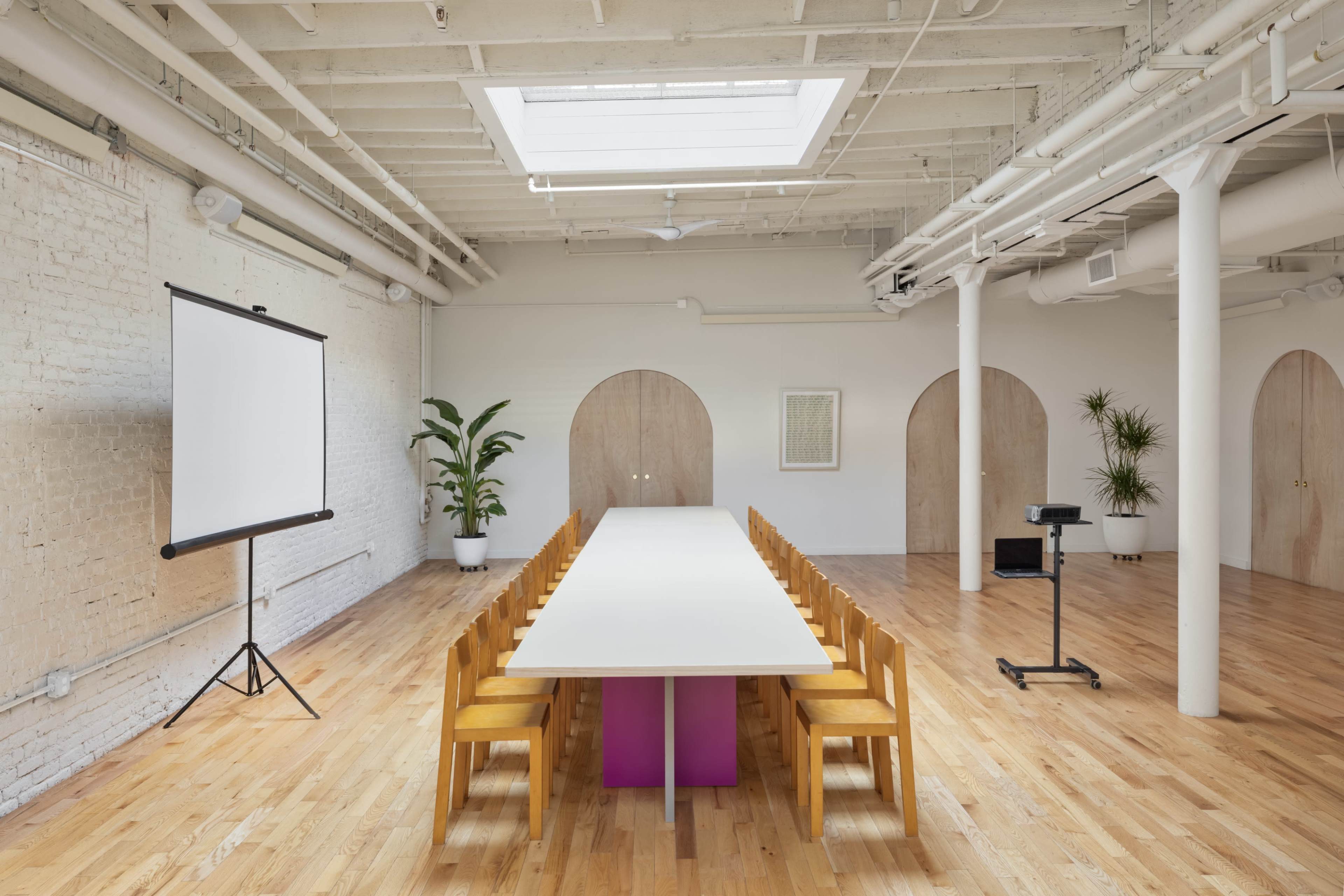 A bright meeting room features a long rectangular table surrounded by wooden chairs, with a projector screen and plants against the walls.