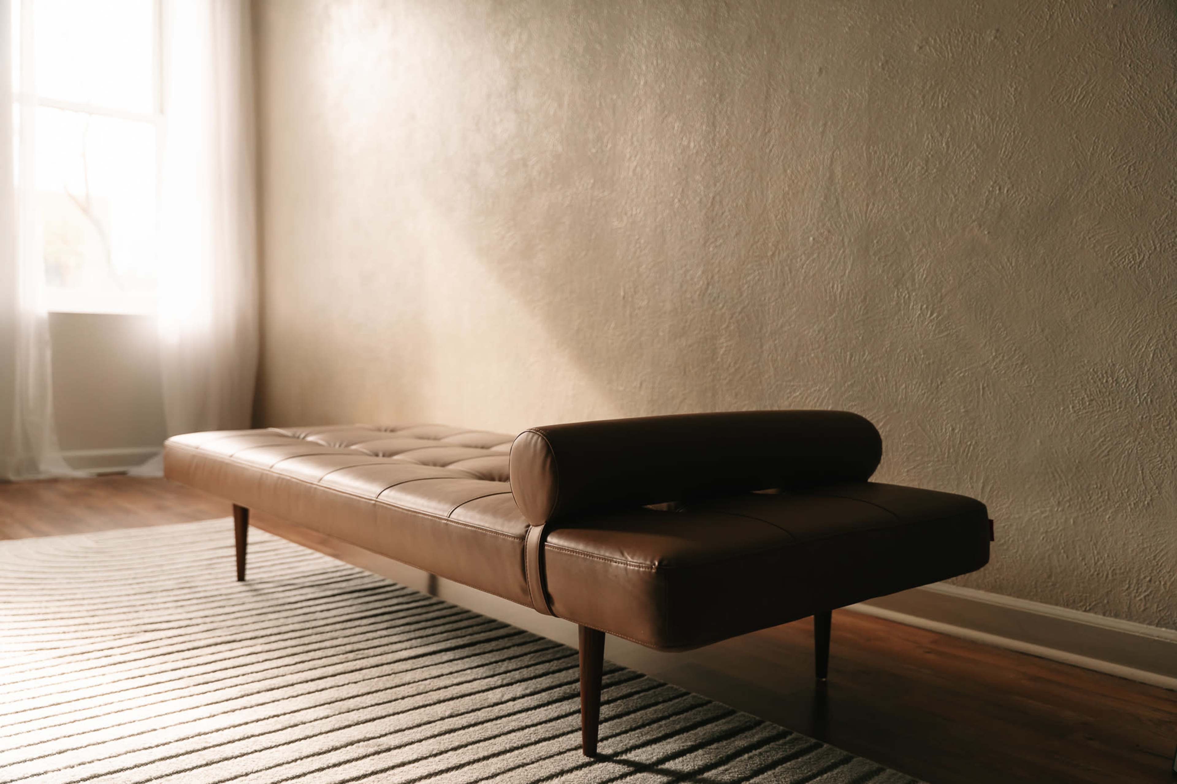 A brown leather daybed is positioned on a striped rug in a well-lit room with textured walls.