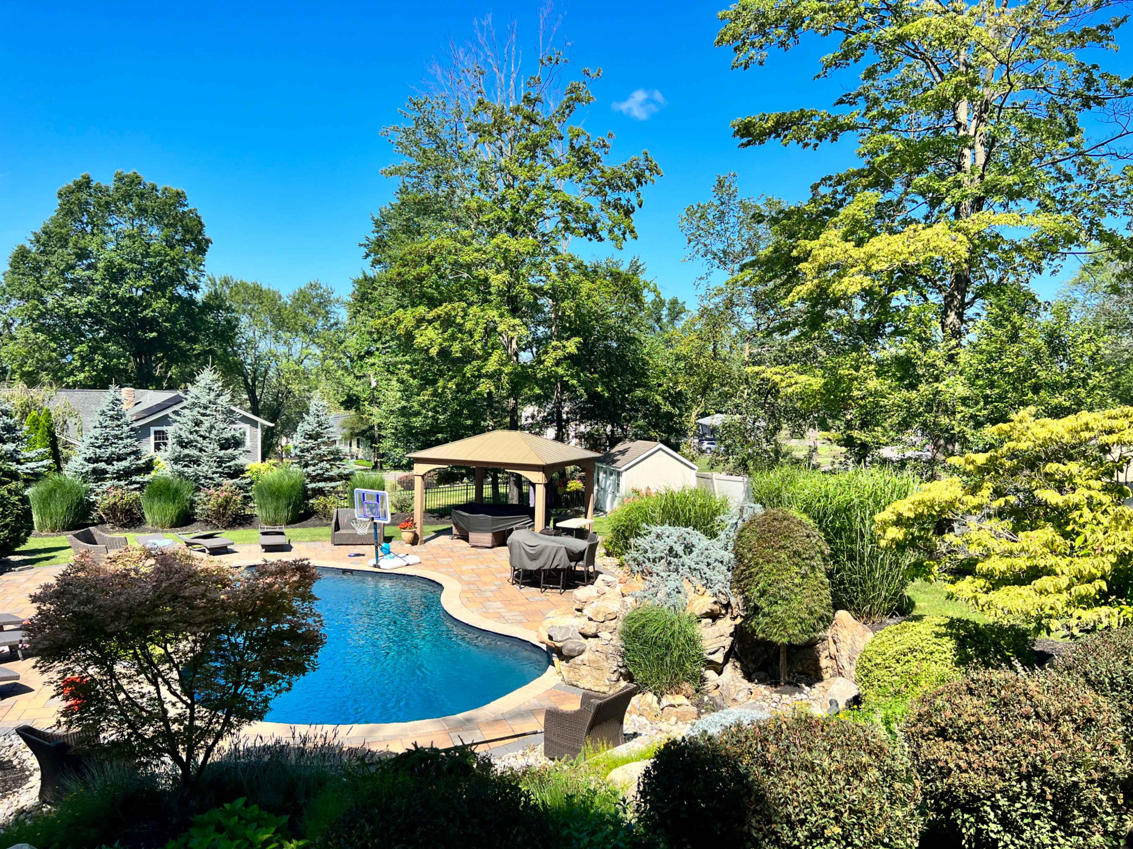 A well-maintained backyard features a swimming pool surrounded by lounge chairs, a gazebo, and landscaped greenery under a clear blue sky.