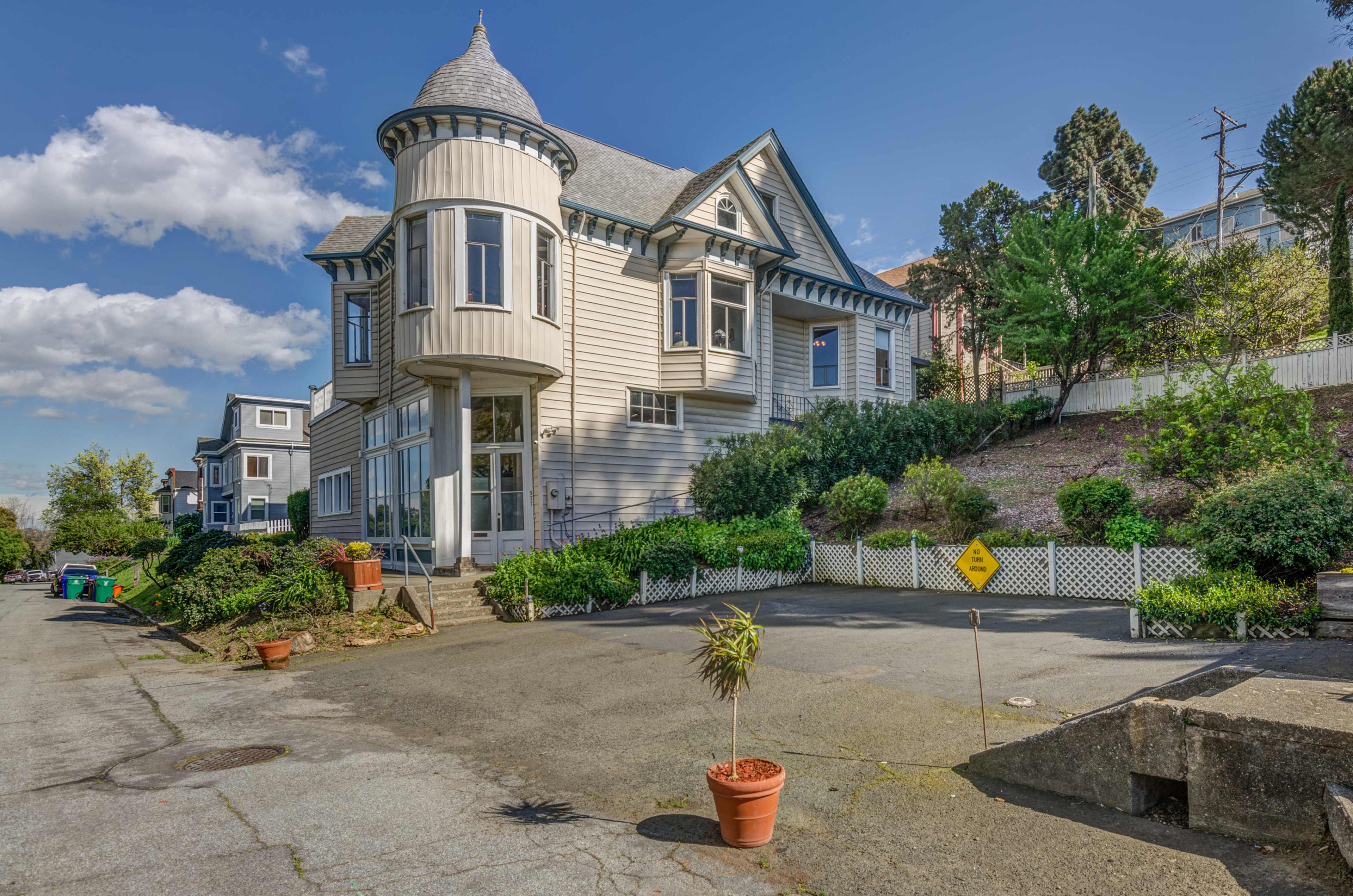 A large, Victorian-style house with a turret, situated on a tree-lined street with neighboring homes and a planted area in the foreground.