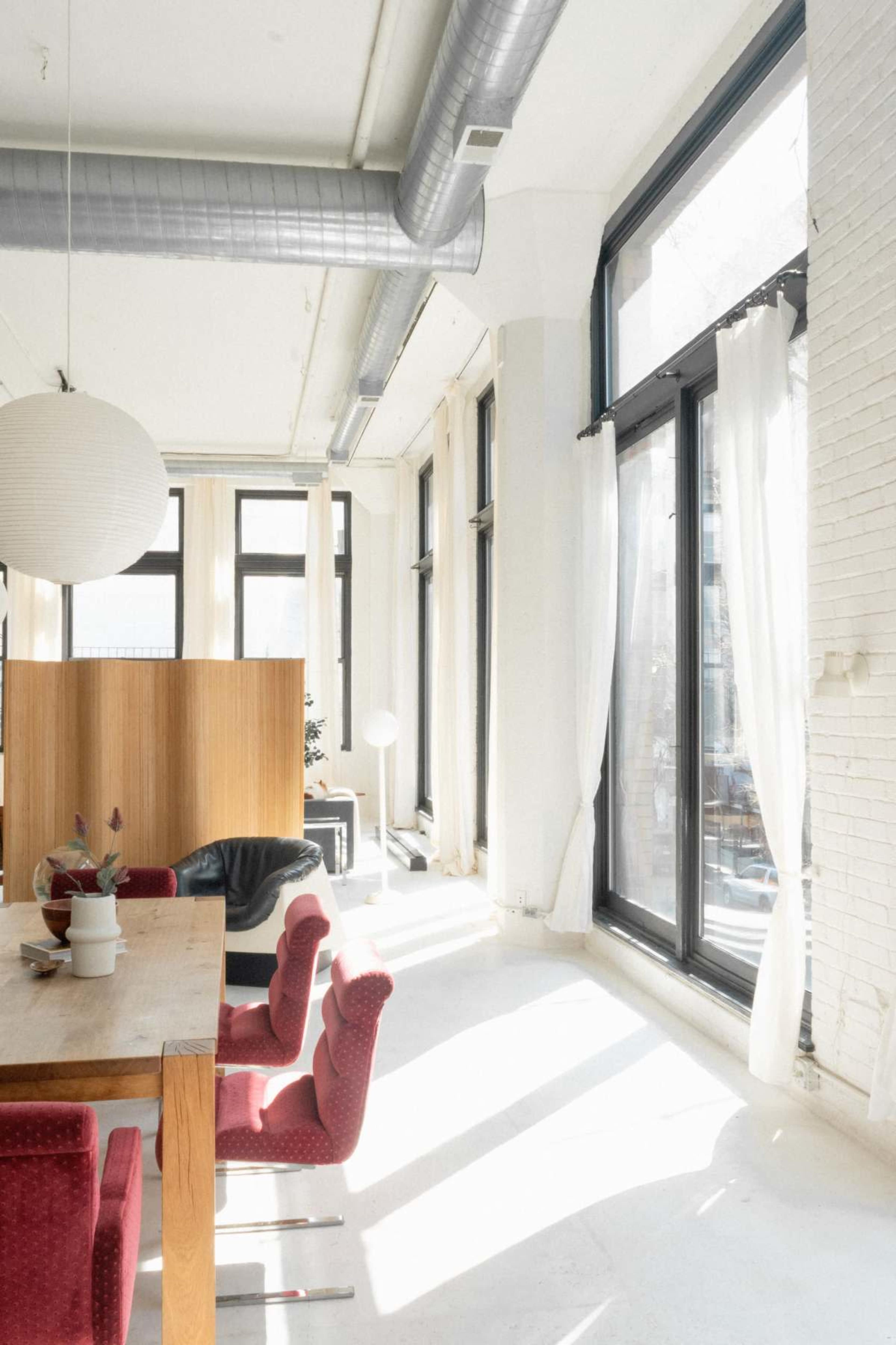A dining area features a wooden table surrounded by red chairs, large windows allowing natural light, and sheer curtains.