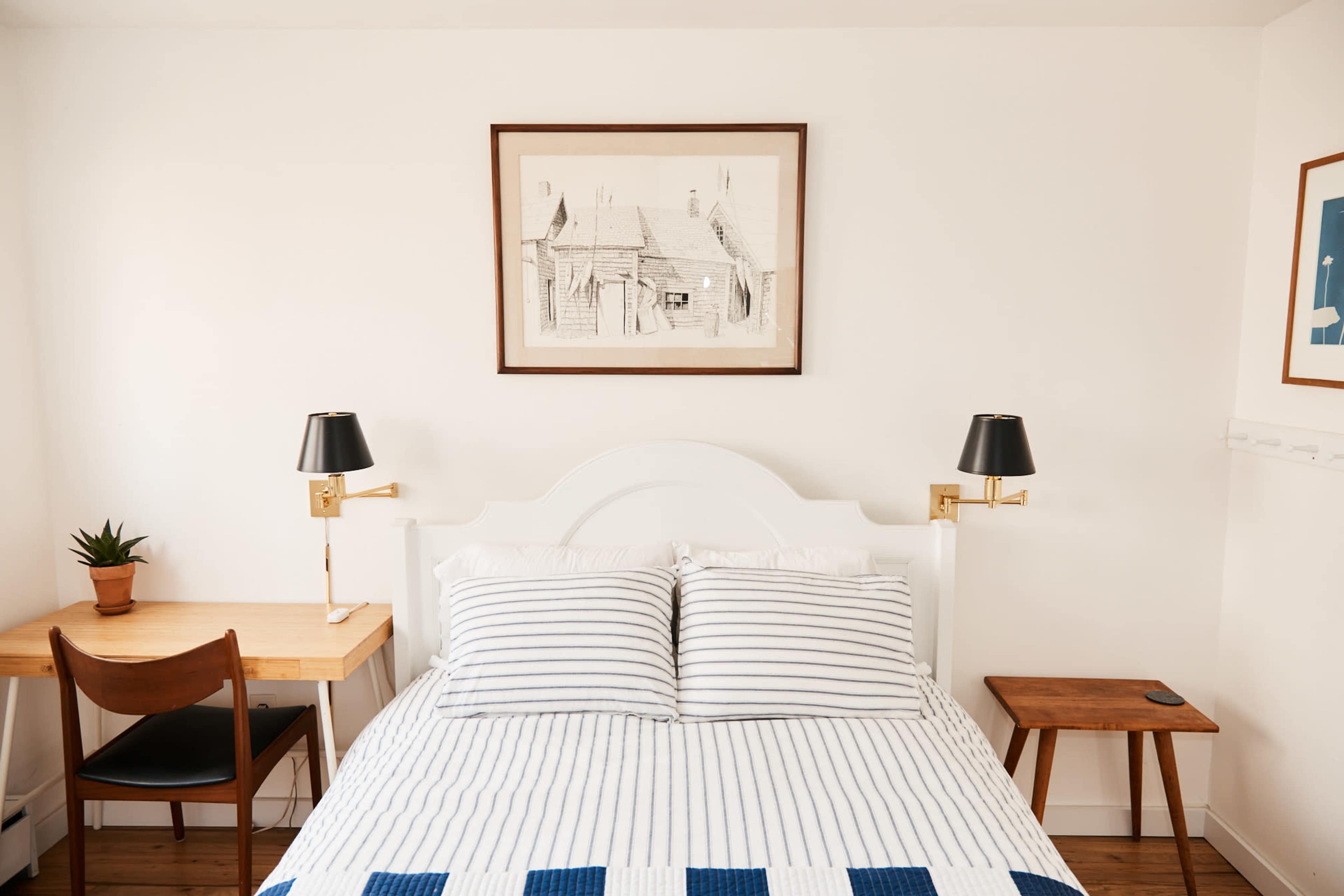 A minimalistic bedroom featuring a bed with striped linens, a framed drawing on the wall, a small desk with a chair, and two wall-mounted lamps.