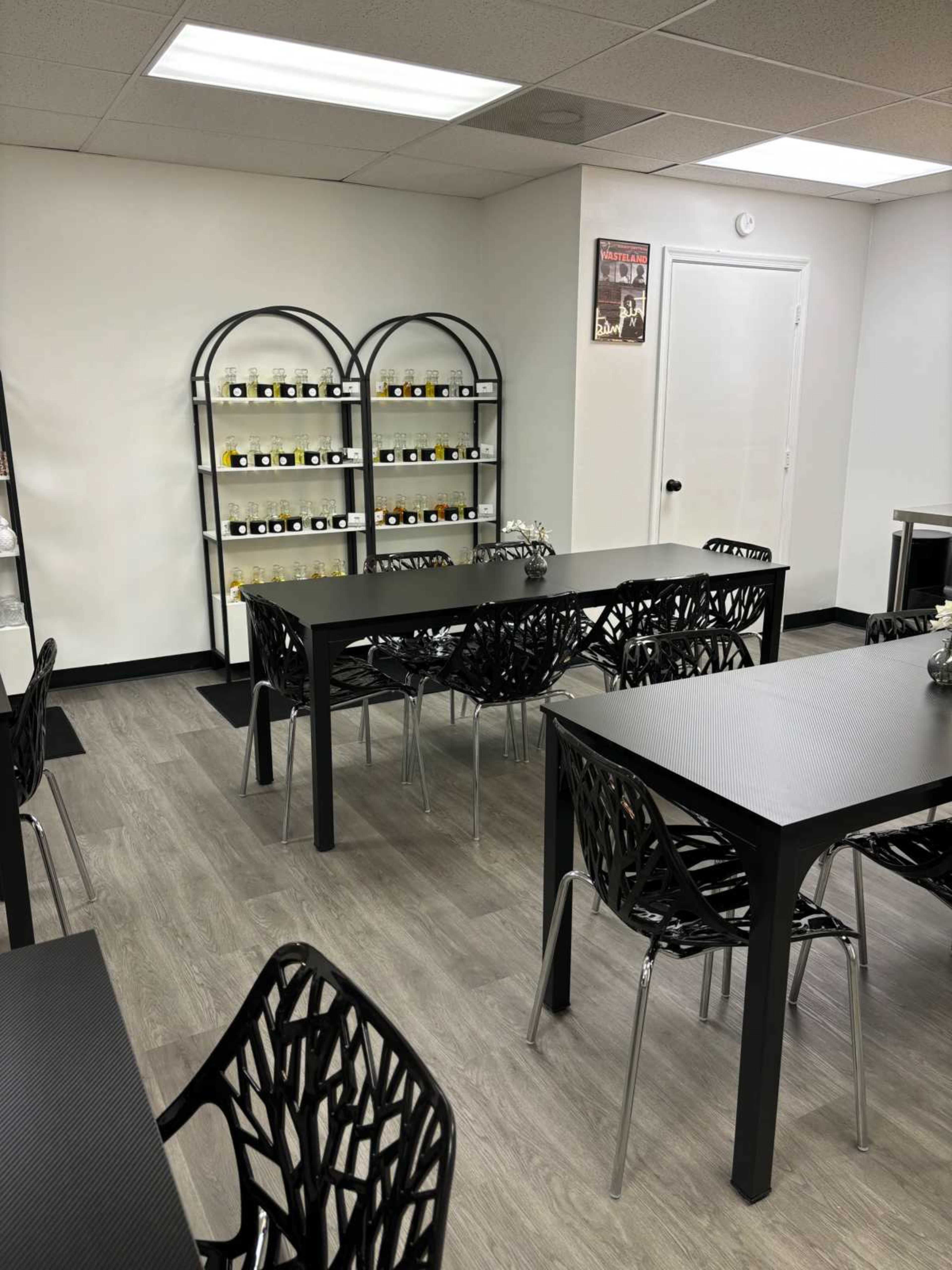 A modern, minimalist classroom or meeting space with black tables, clear plastic chairs, and shelves displaying jars.
