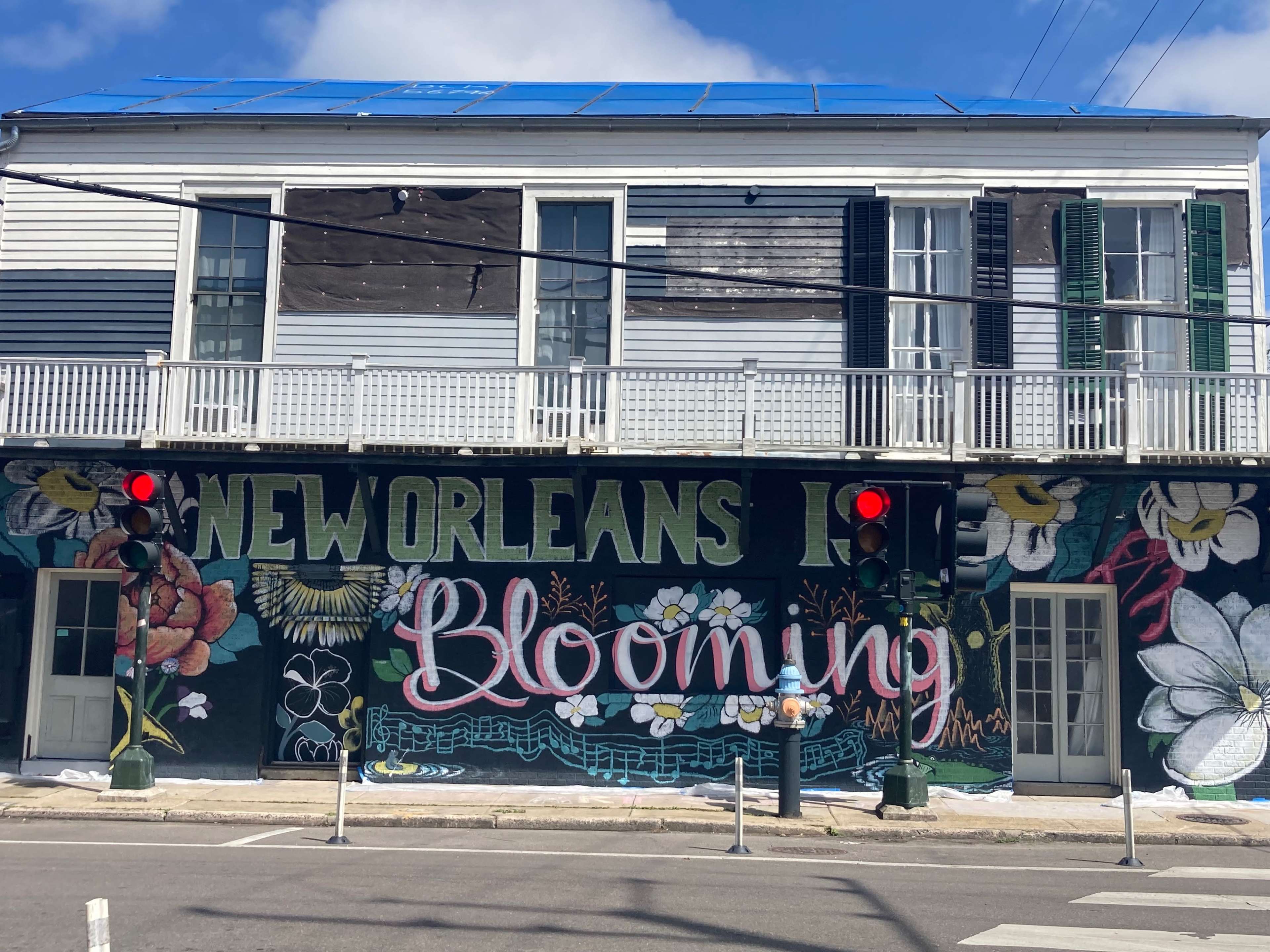 The image shows a vibrant mural on a building in New Orleans with the text "New Orleans Is Blooming" surrounded by colorful floral designs.