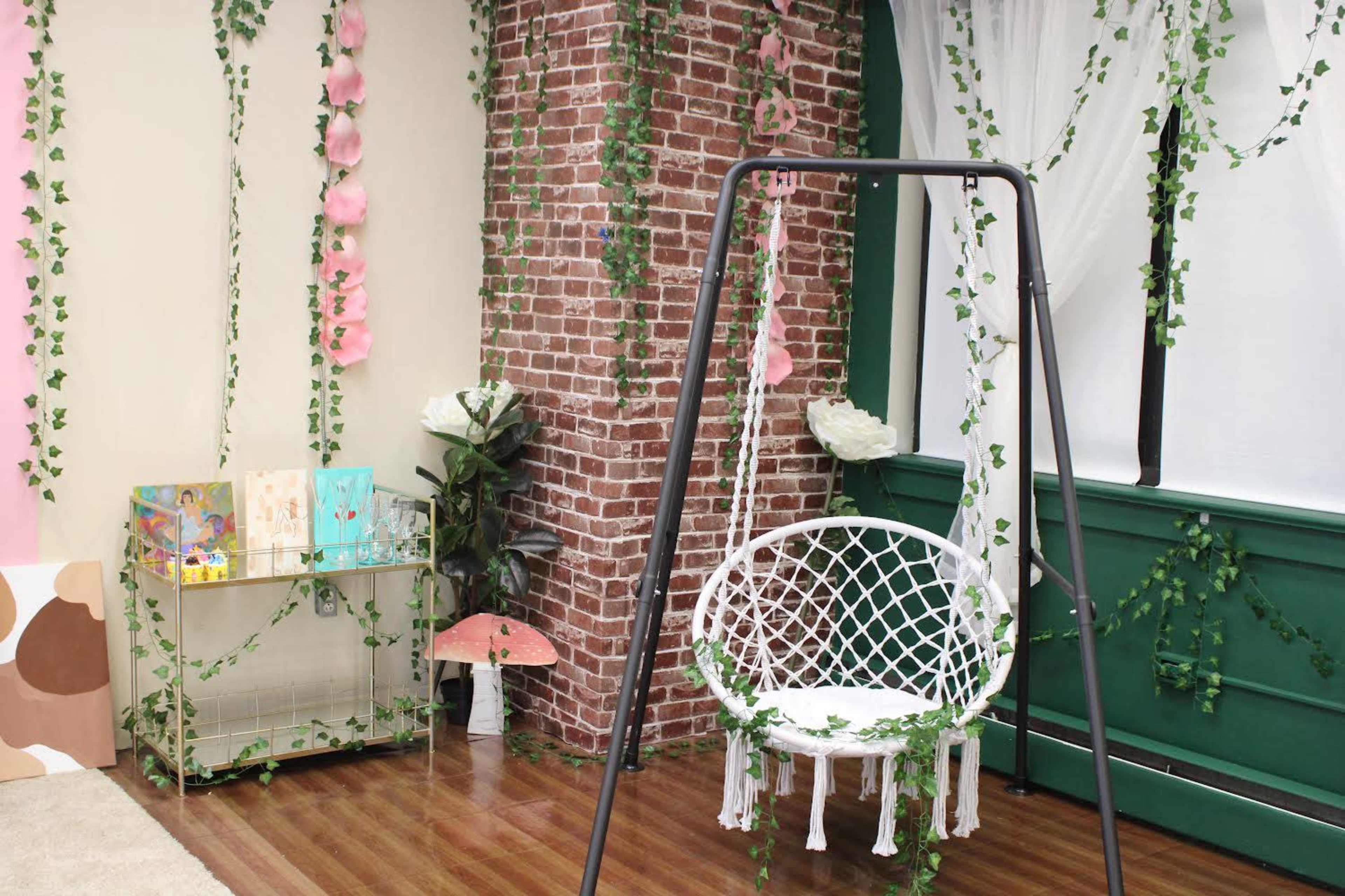 The scene features a swing chair surrounded by greenery and brick walls, with decorative plants and art displayed nearby.