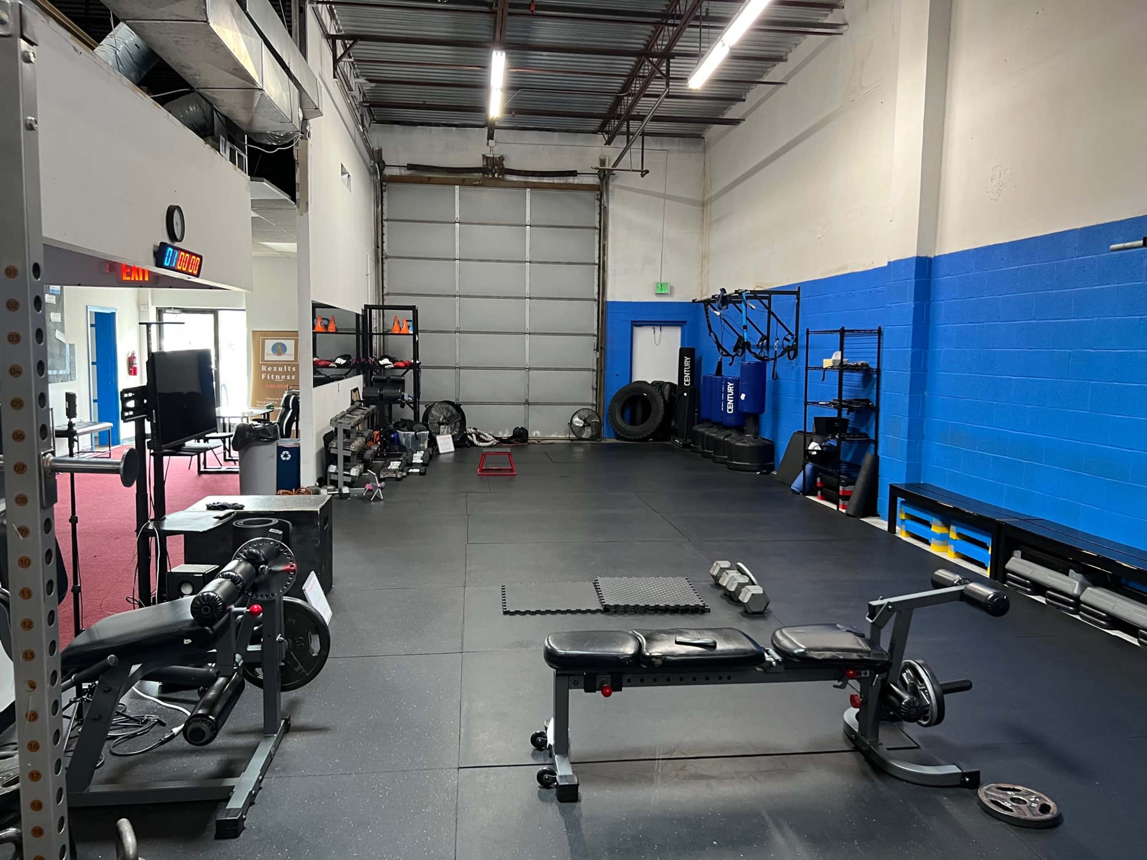 An empty gym room with various fitness equipment, including a weight bench, exercise machines, and mats, positioned against blue and gray walls.