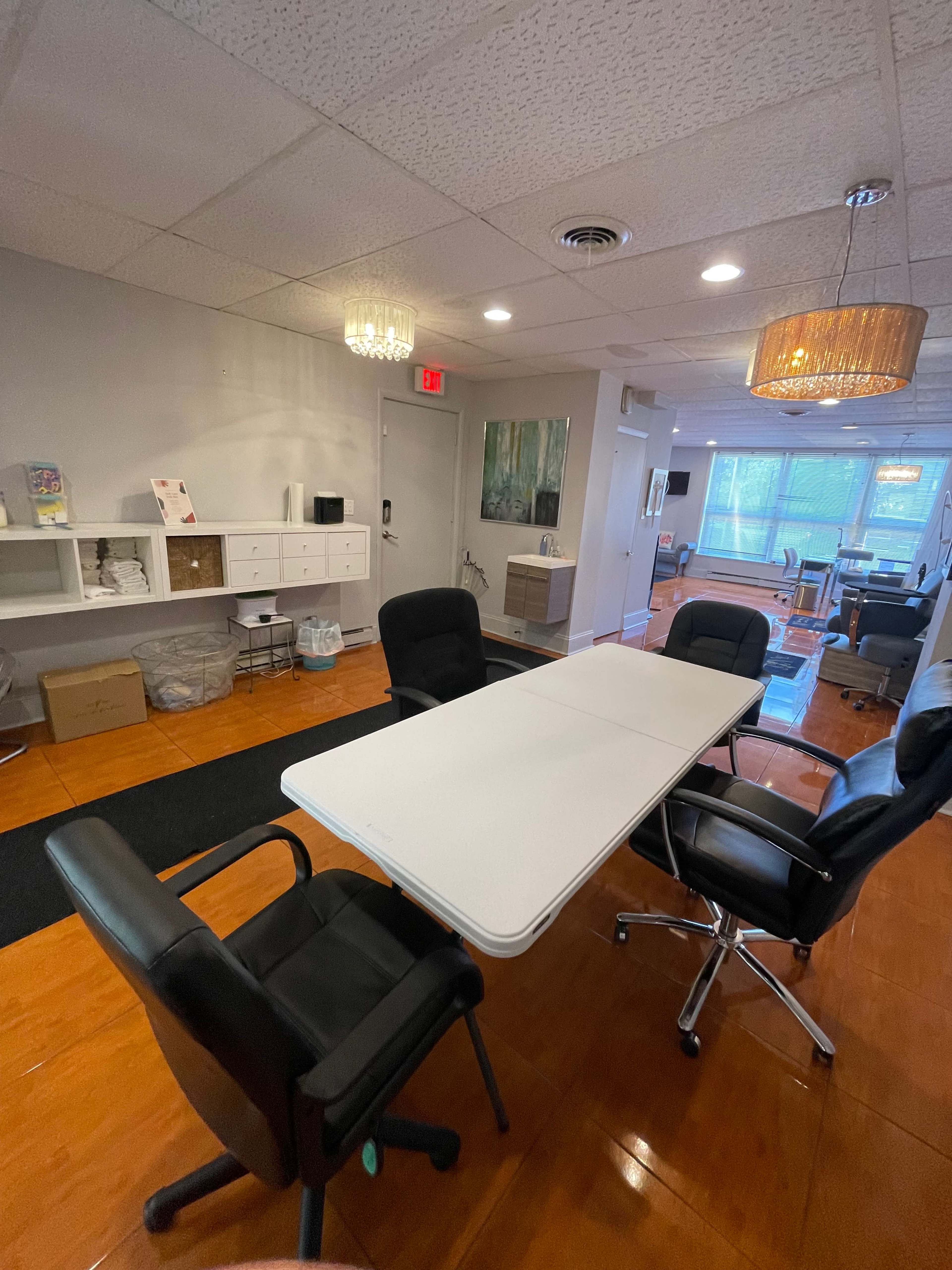 A well-lit office space featuring a long table surrounded by chairs, with shelves and a seating area in the background.