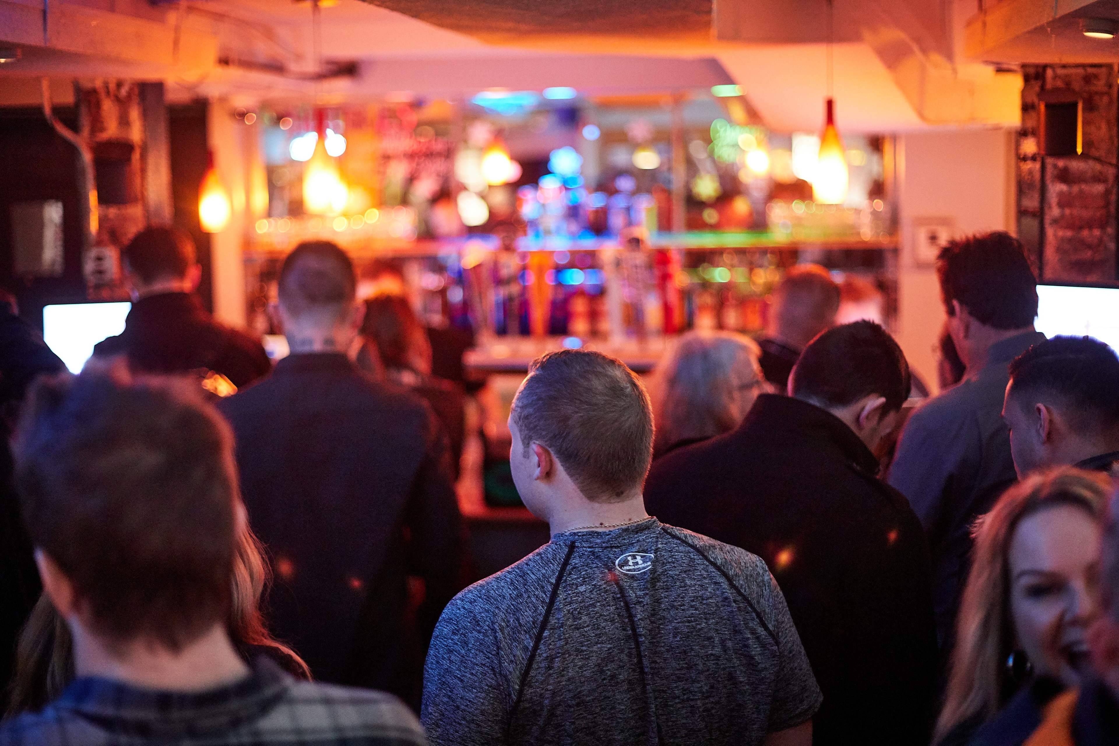 A group of people stands with their backs to the camera in a dimly lit bar, with a brightly lit bar counter visible in the background.