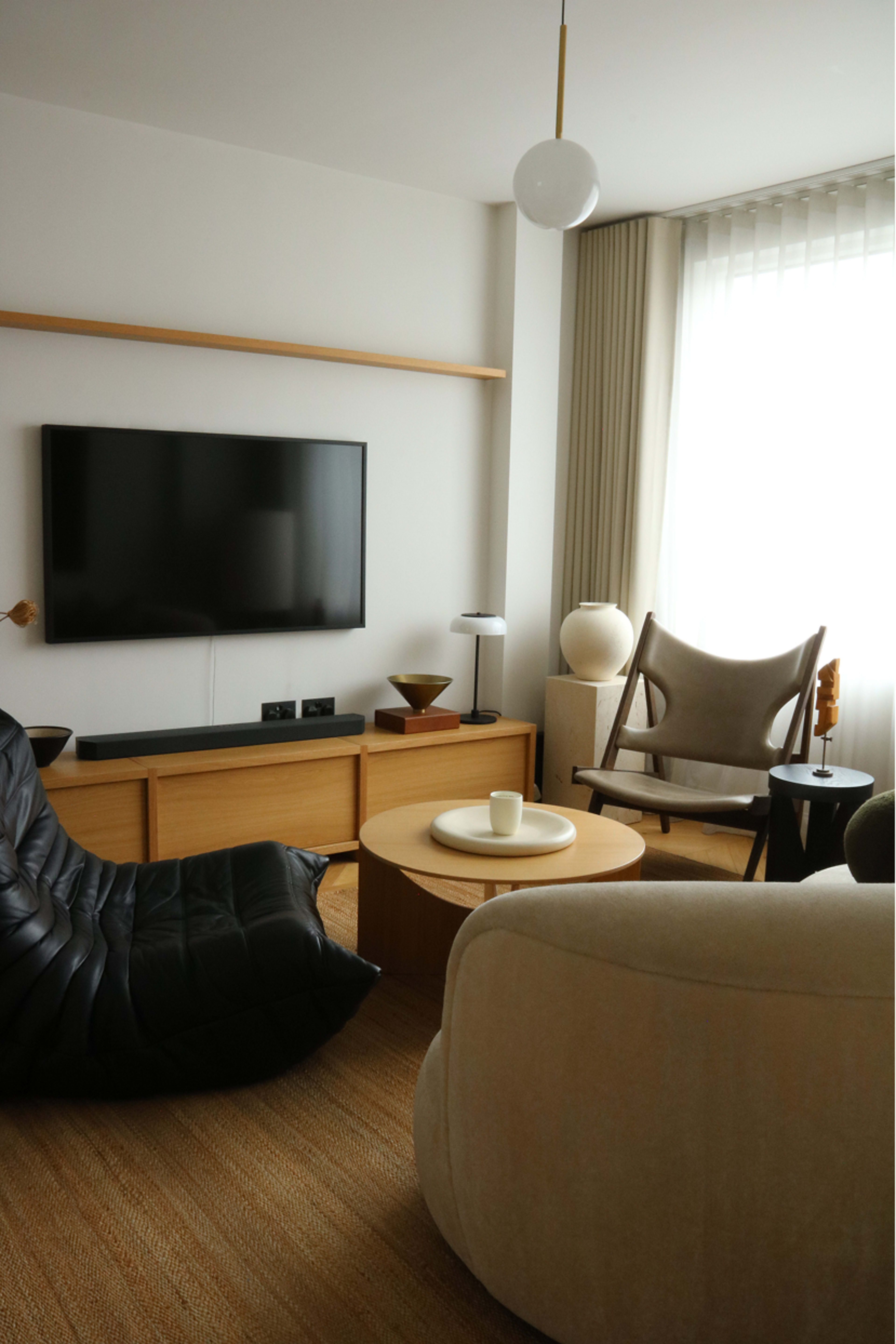 The image shows a modern living room featuring a black bean bag chair, a cozy cream-colored armchair, a round wooden coffee table, and a wall-mounted television against a light-colored wall.
