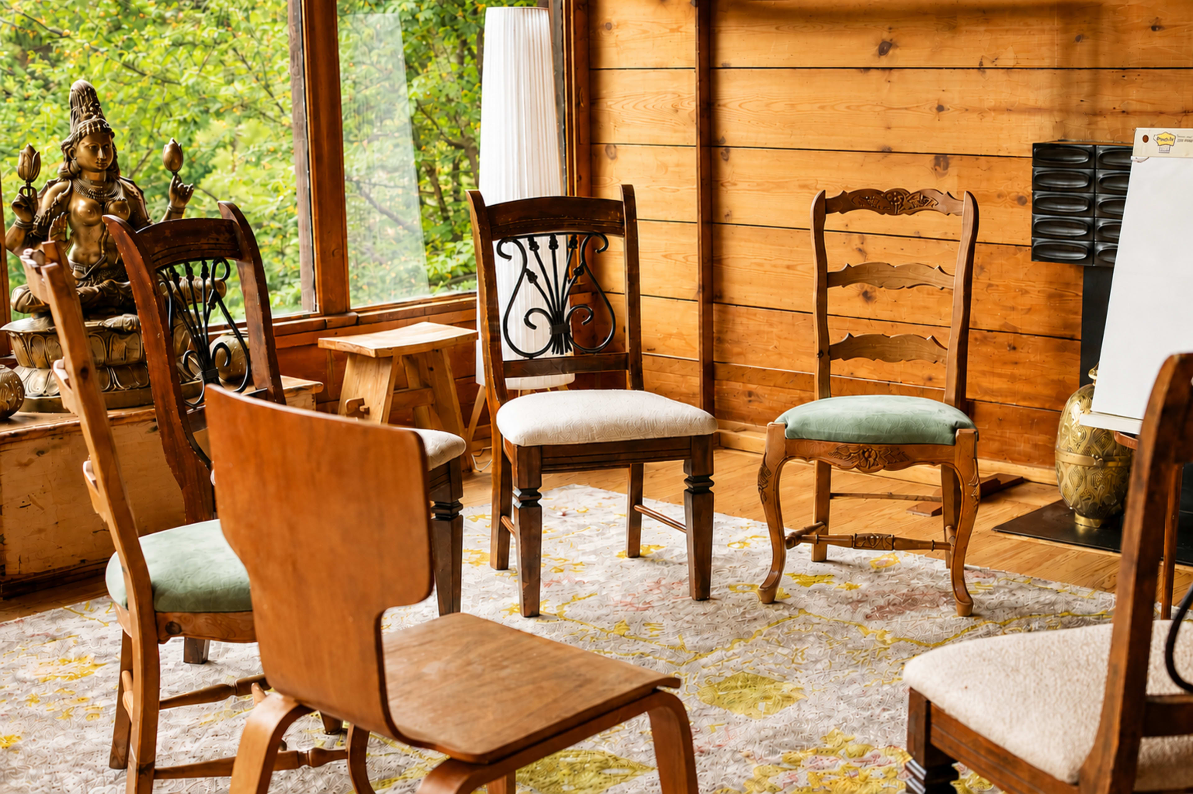 A set of wooden chairs is arranged in a circle on a patterned rug inside a cozy, wooden-paneled room with a decorative statue in the corner.