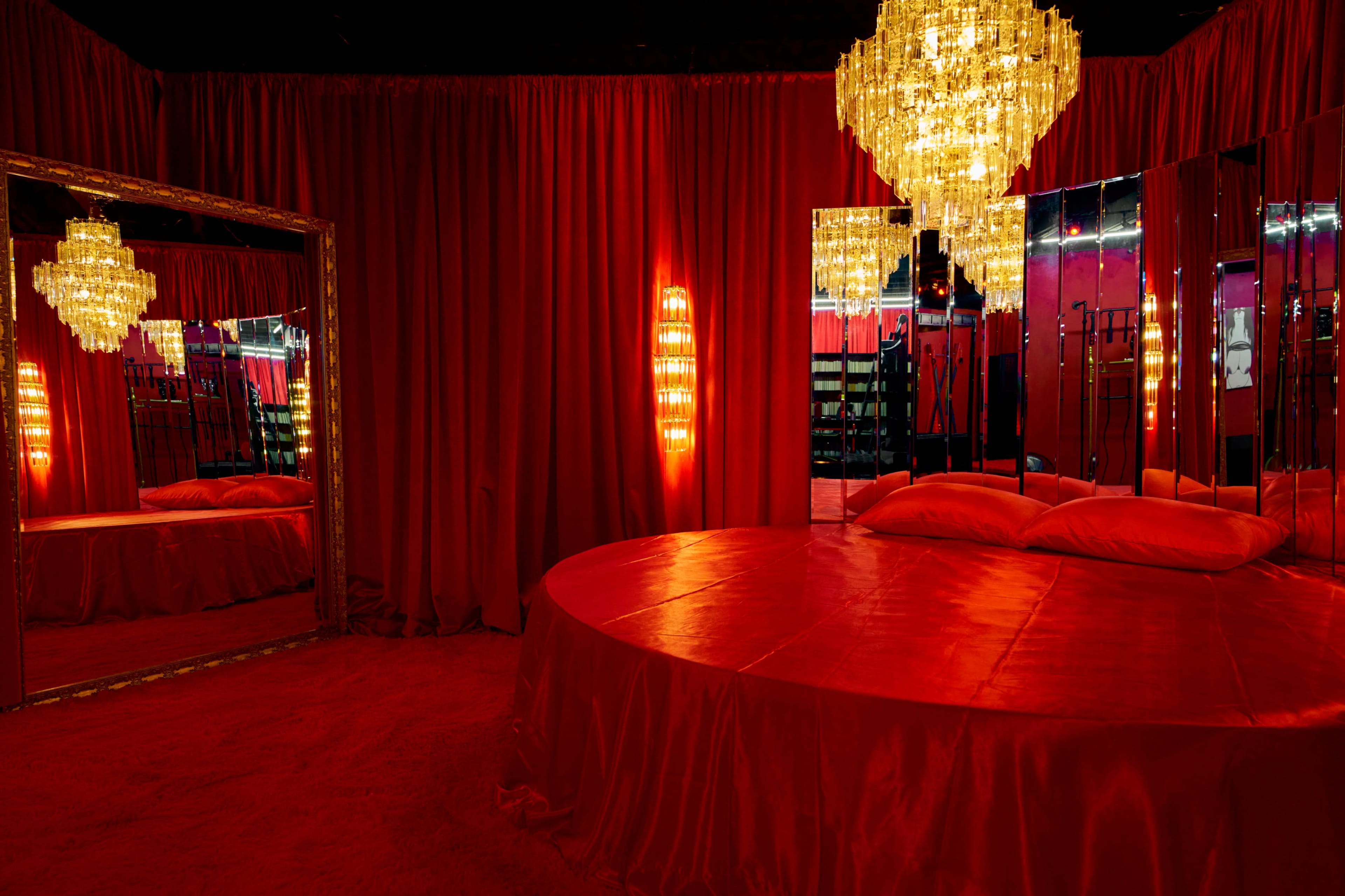 The scene features a circular red bed surrounded by red curtains, mirrors, and a chandelier.