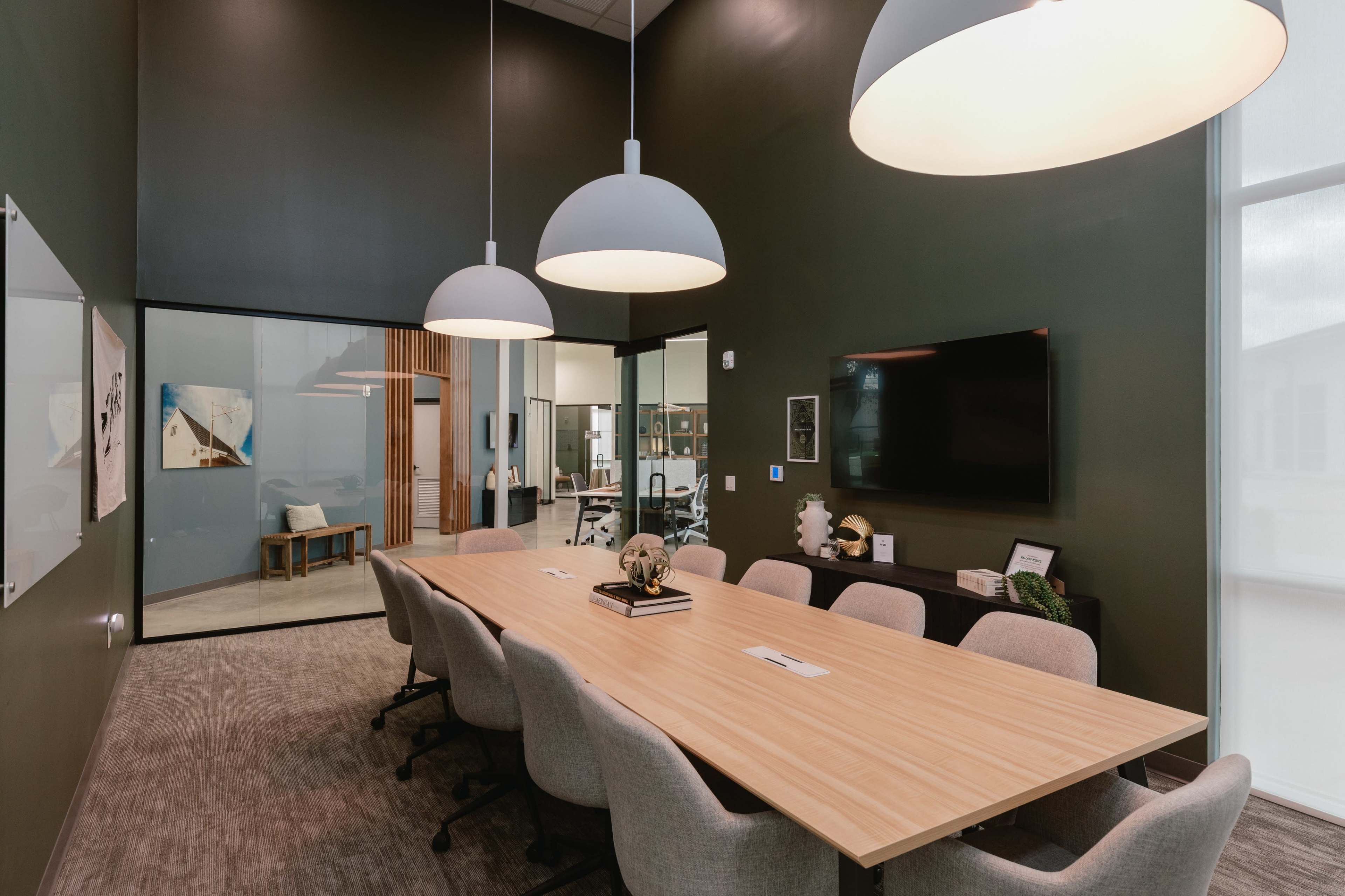 A conference room features a long wooden table surrounded by several chairs and is illuminated by three large pendant lights.