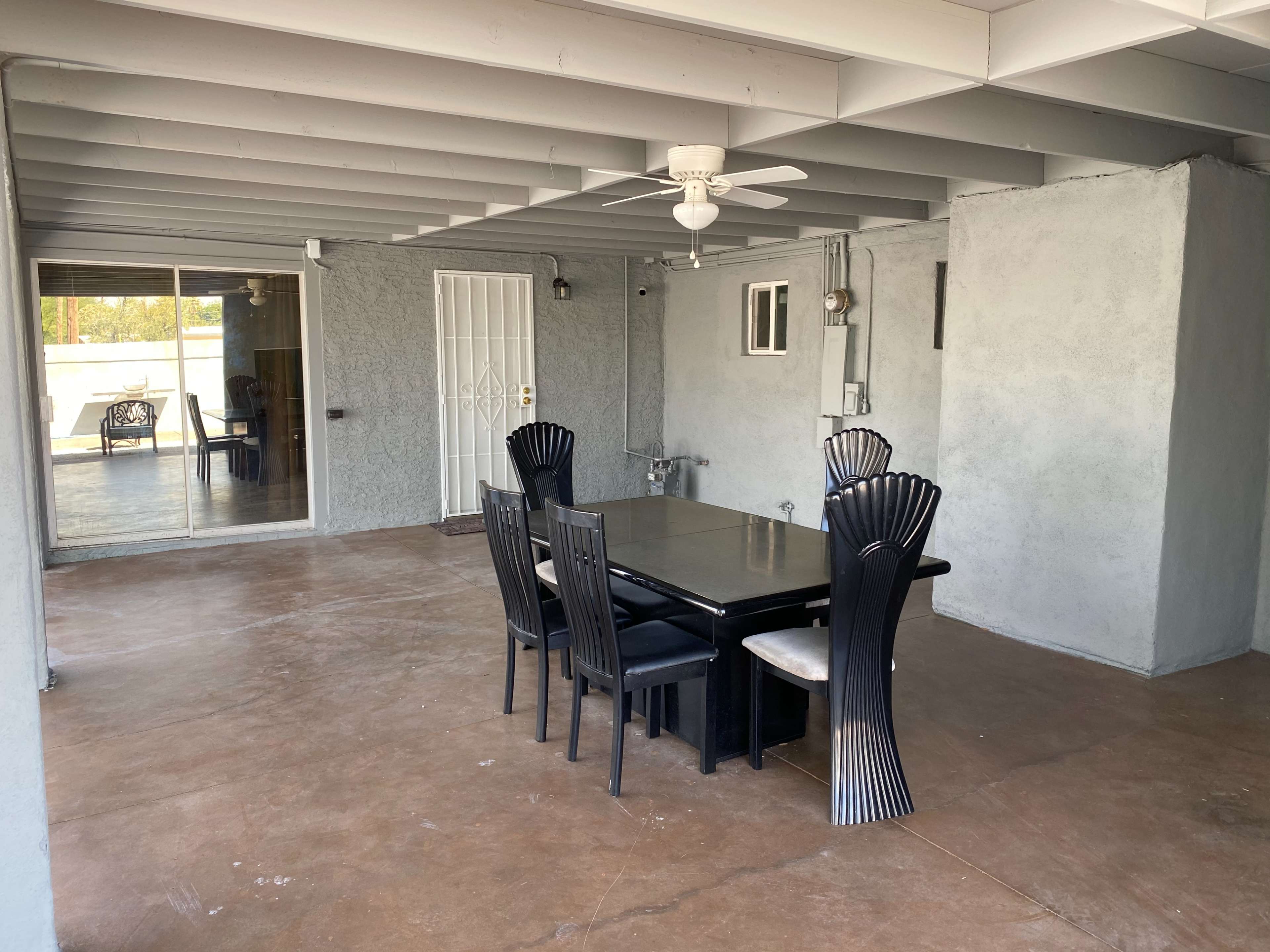 A covered patio featuring a black dining table with six chairs and a ceiling fan, with sliding glass doors leading to an indoor space in the background.