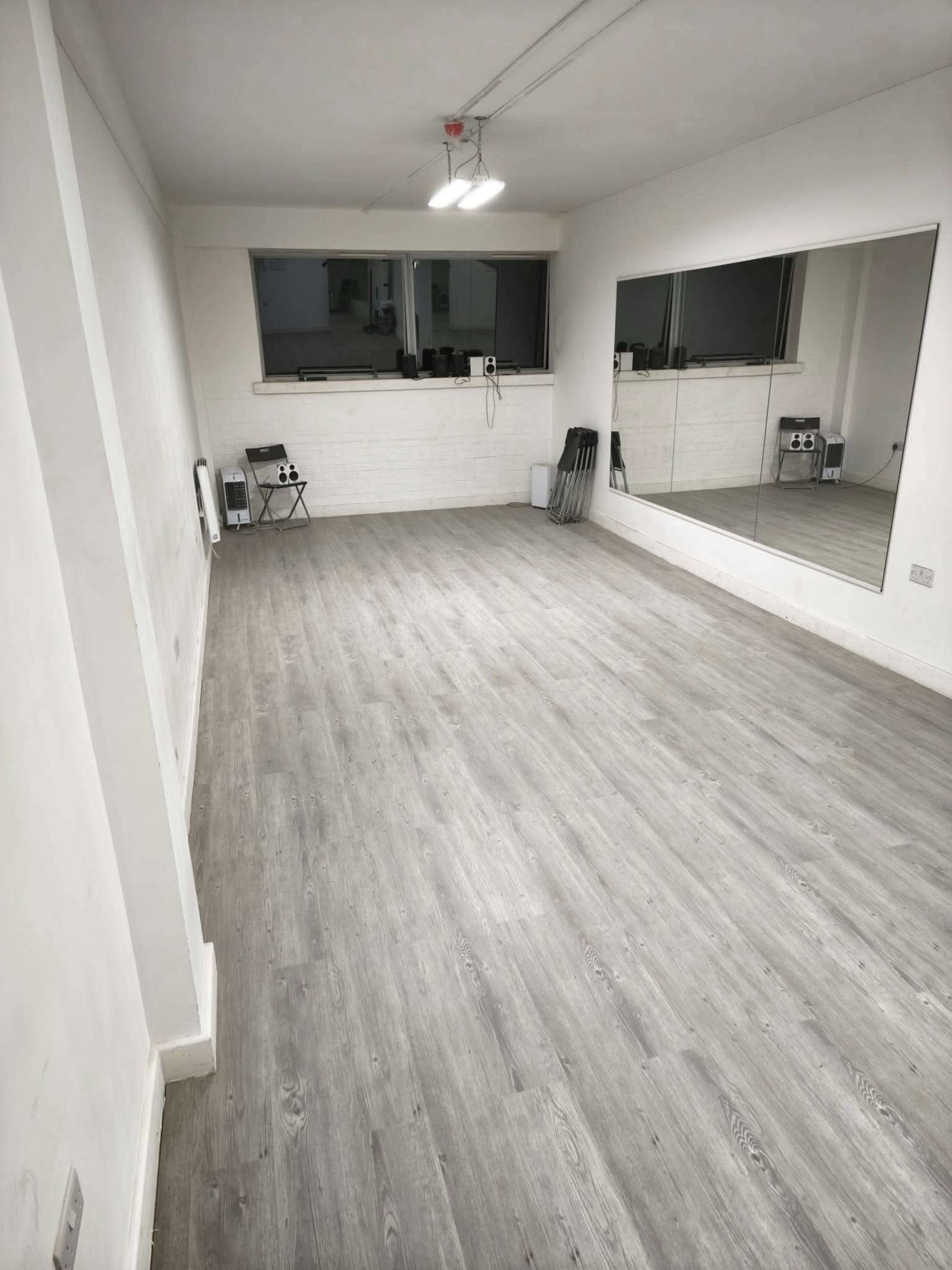 The image shows a spacious, empty room with laminate flooring, a large mirror on one wall, and minimal furnishings.
