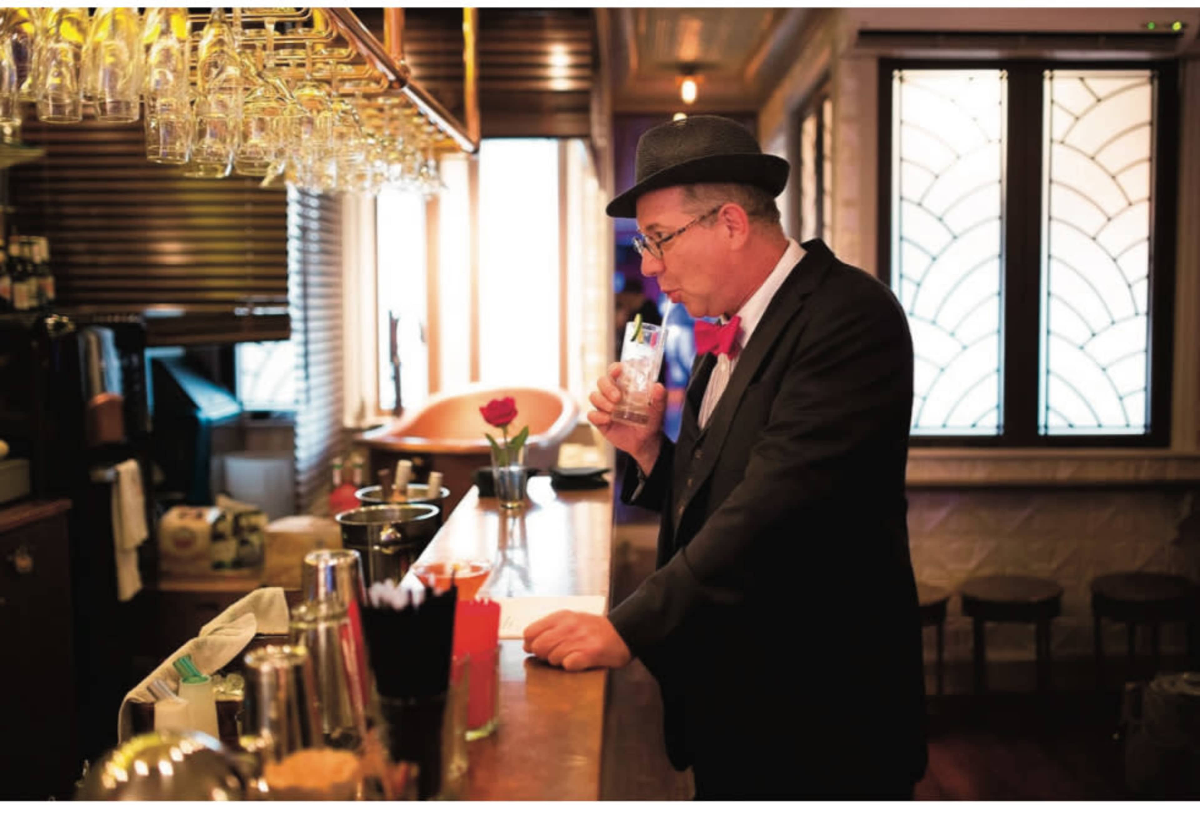 A man in a suit and hat drinks from a glass at a bar with various utensils and decor in the background.