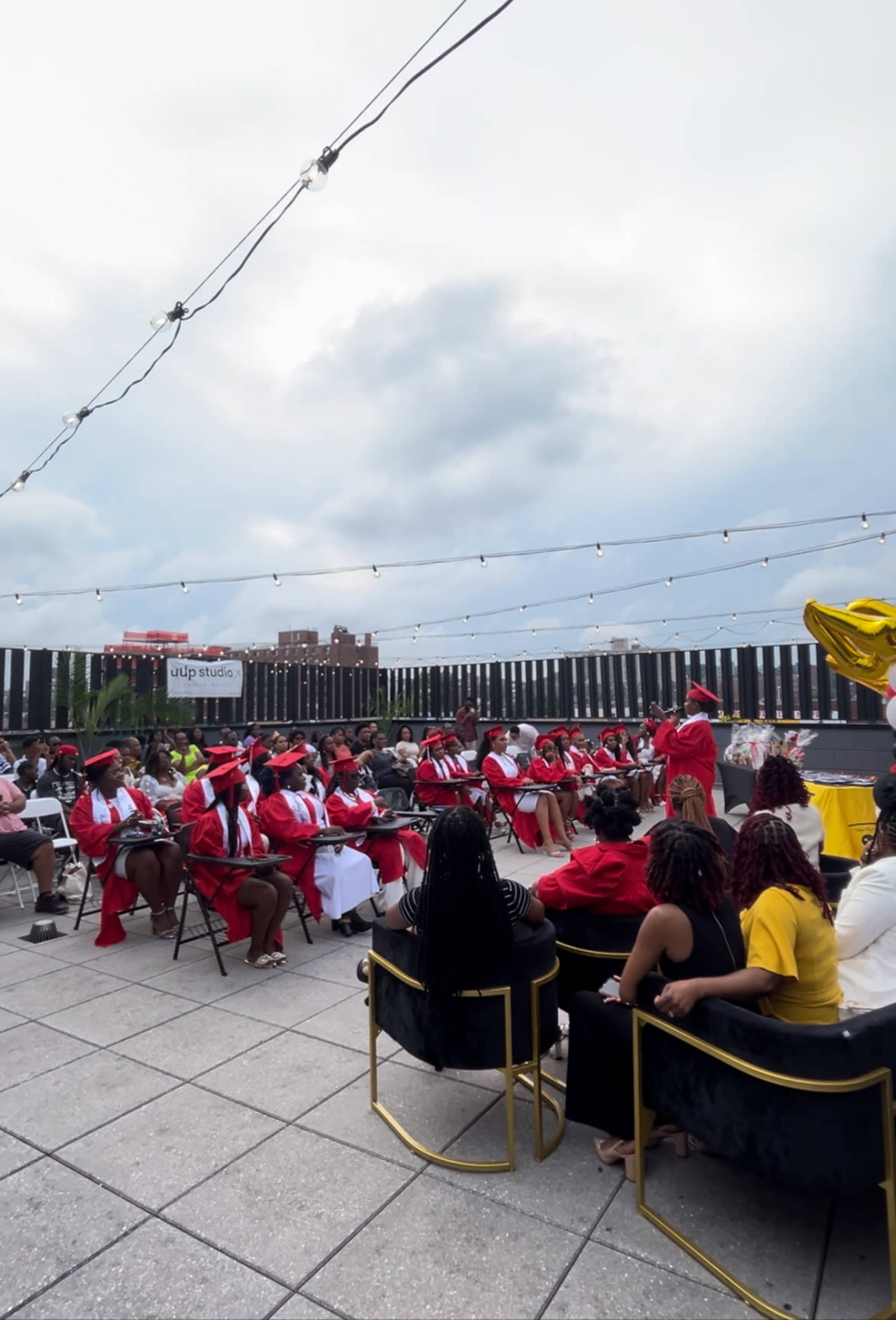 A graduation ceremony takes place outdoors on a rooftop, with attendees seated and wearing red caps and gowns under a cloudy sky.